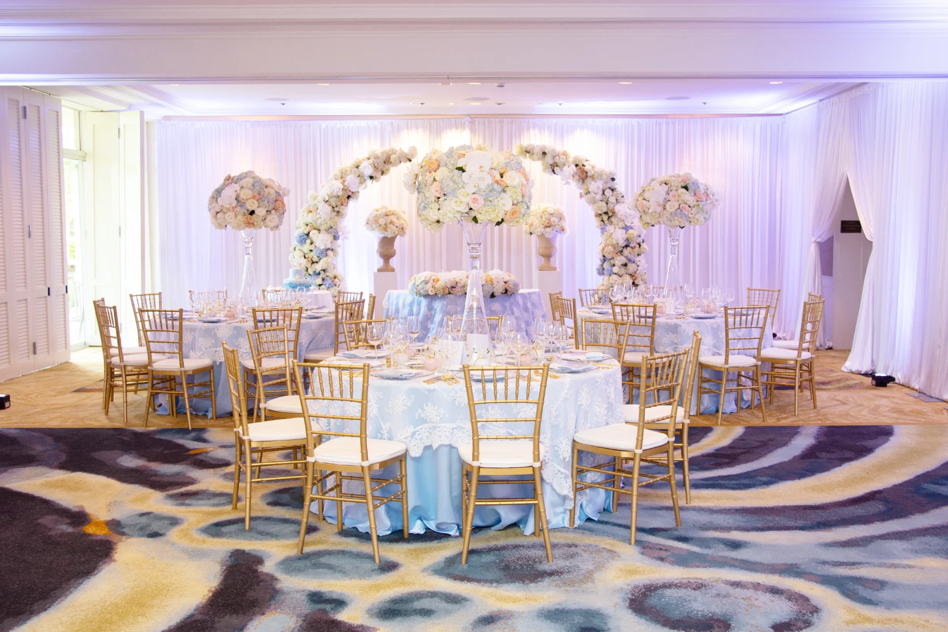 Elegant indoor wedding setup at Four Seasons Oahu with blue and white floral arrangements, gold chairs, and light blue tablecloths.