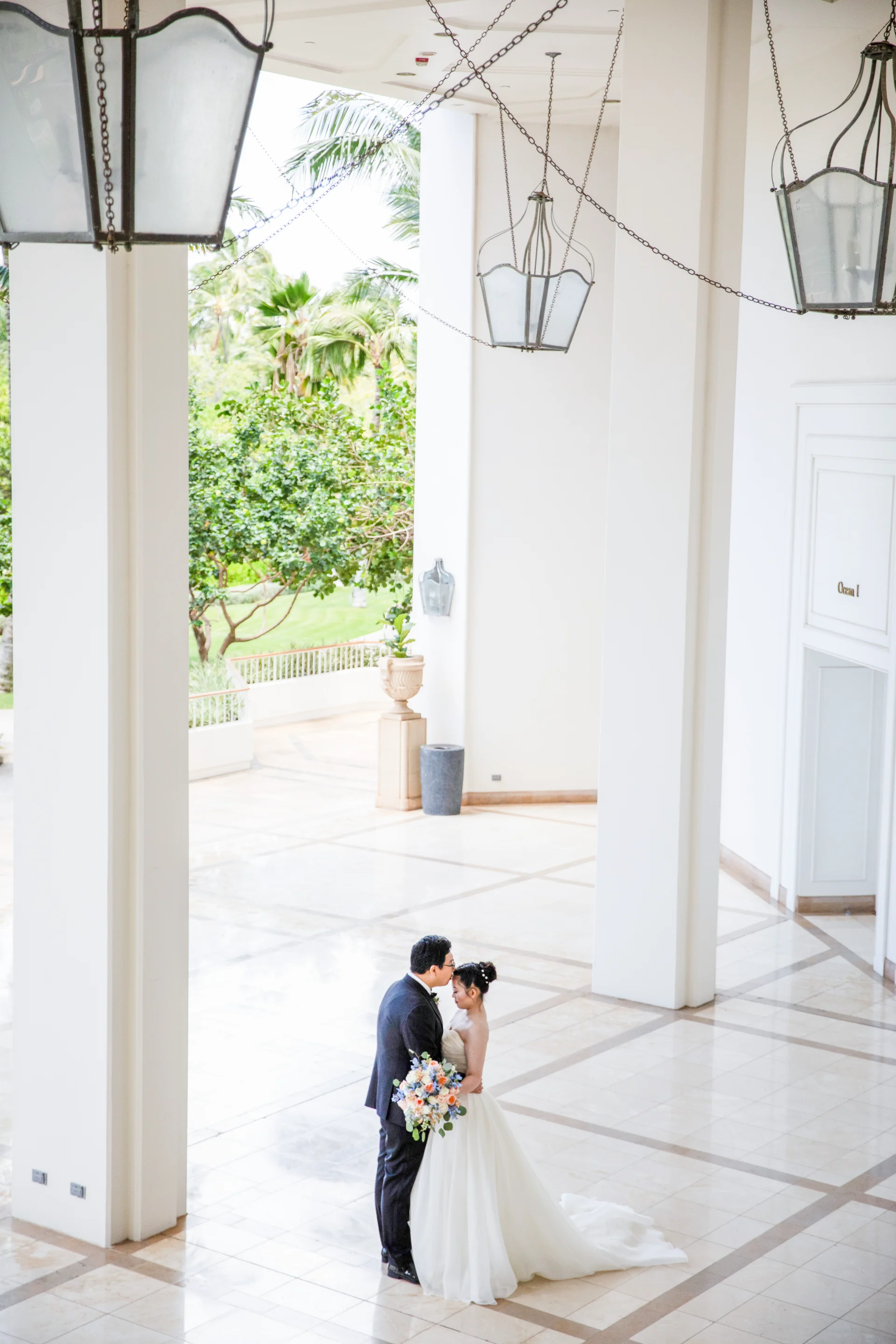 Bride and groom with luxurious bouquet under grand chandeliers at Four Seasons Oahu at Ko'olina, surrounded by greenery.