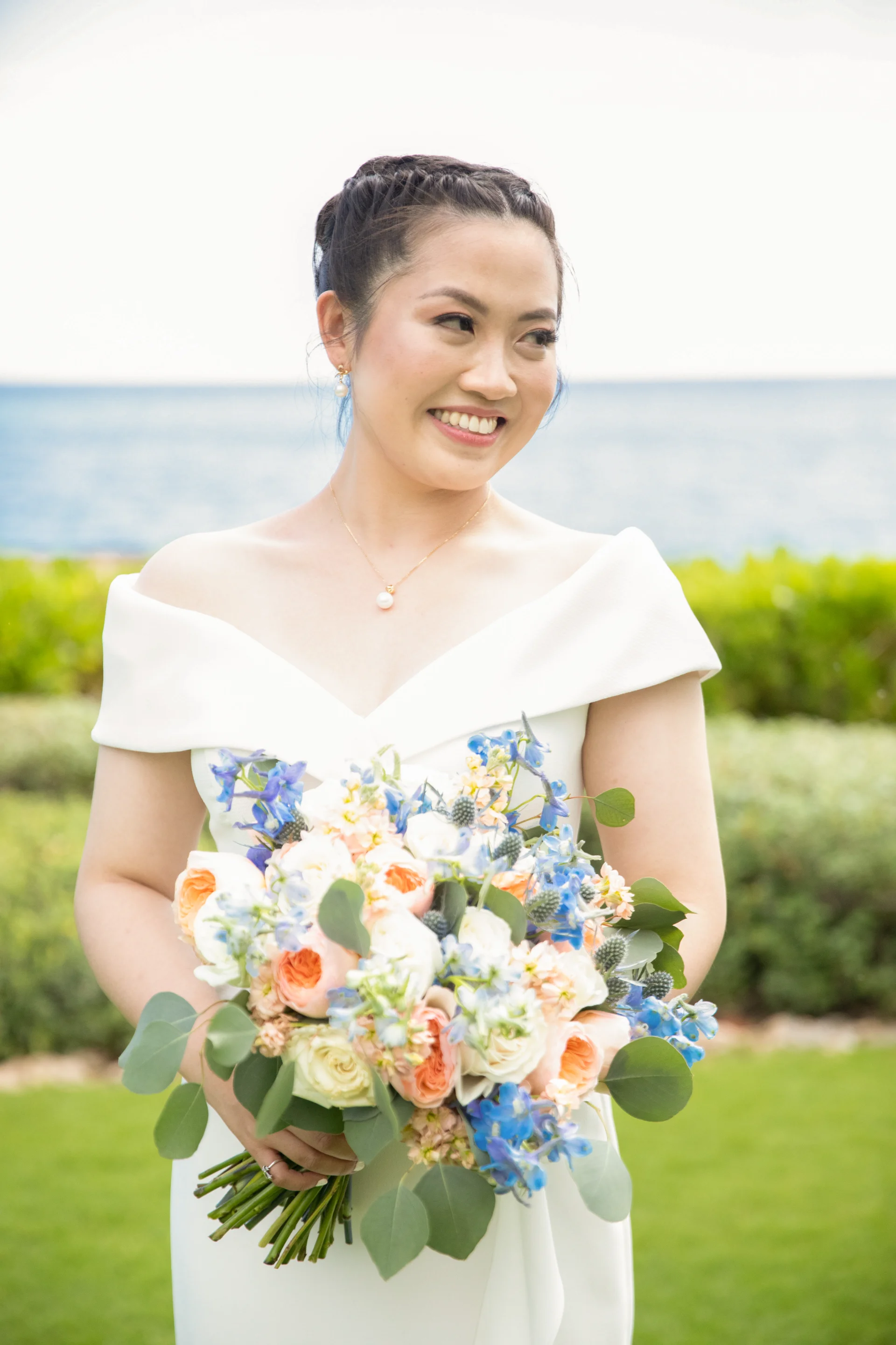 Bride holding a bouquet of peach, white, and blue flowers at Four Seasons Oahu with ocean backdrop.