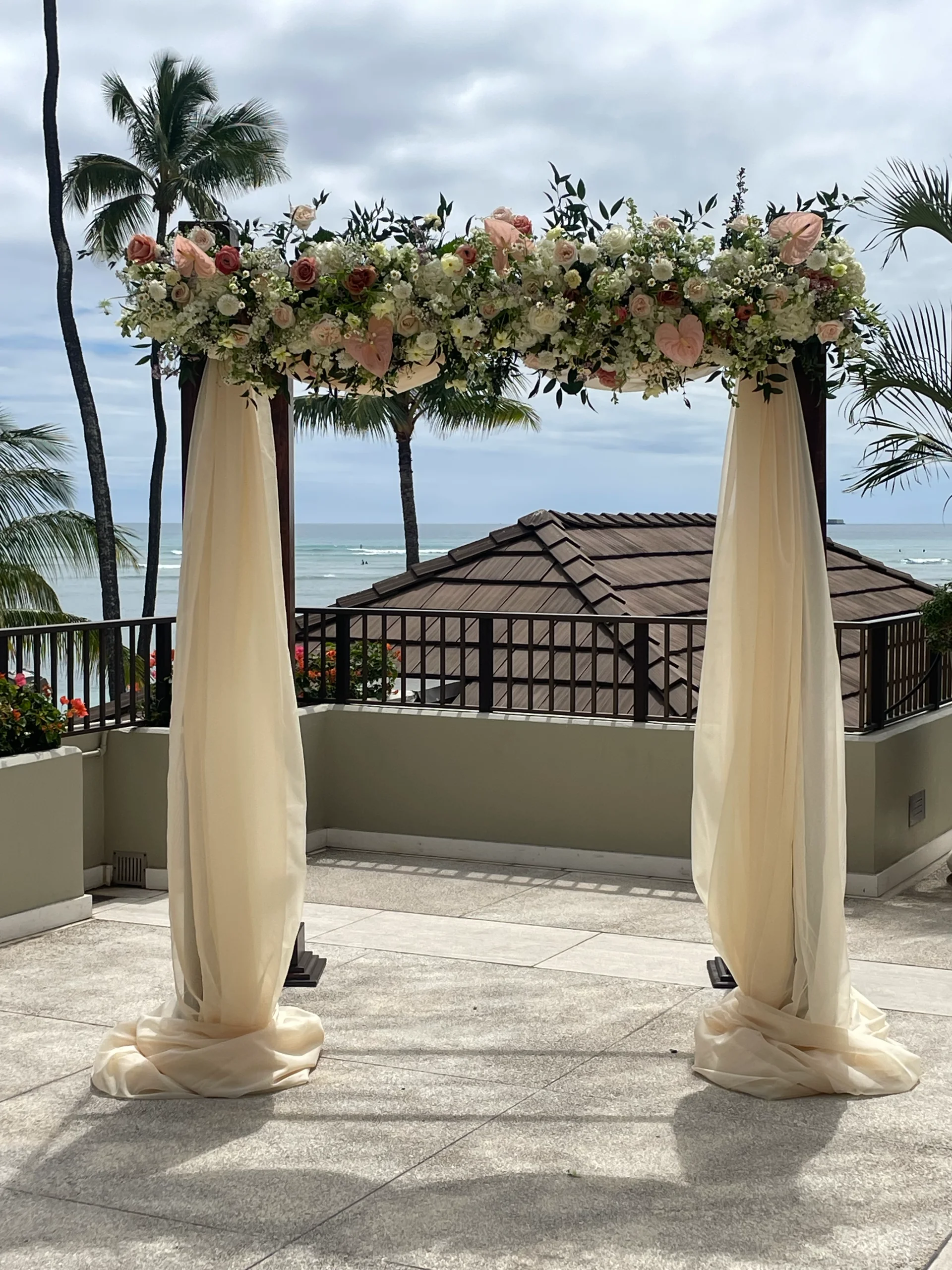 Elegant wedding arch with lush floral arrangement featuring blush roses and greenery, set on a terrace overlooking the ocean in Oʻahu, Hawaii.