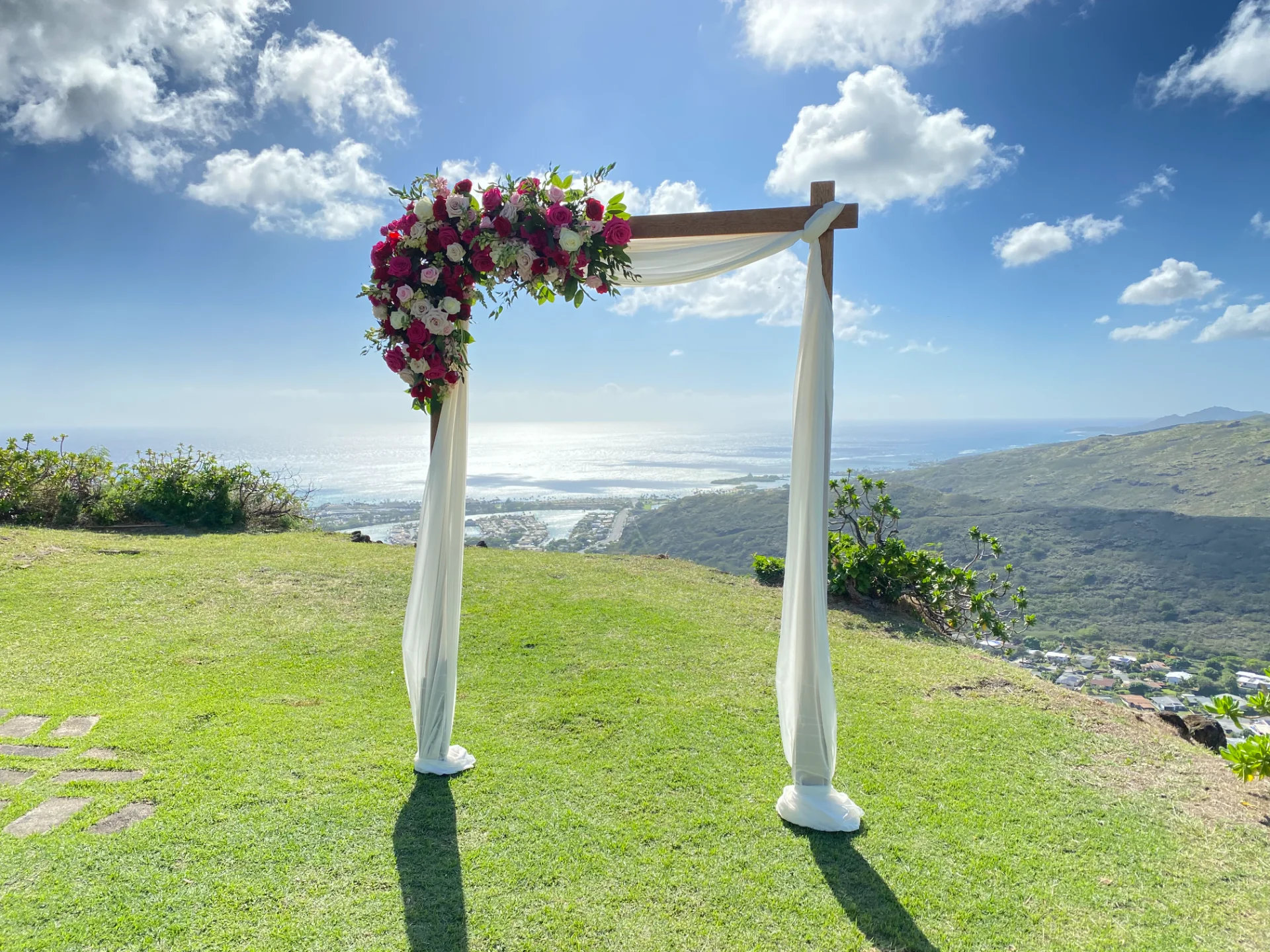 Scenic Oʻahu wedding arch adorned with vibrant red and white florals, overlooking ocean and hills.