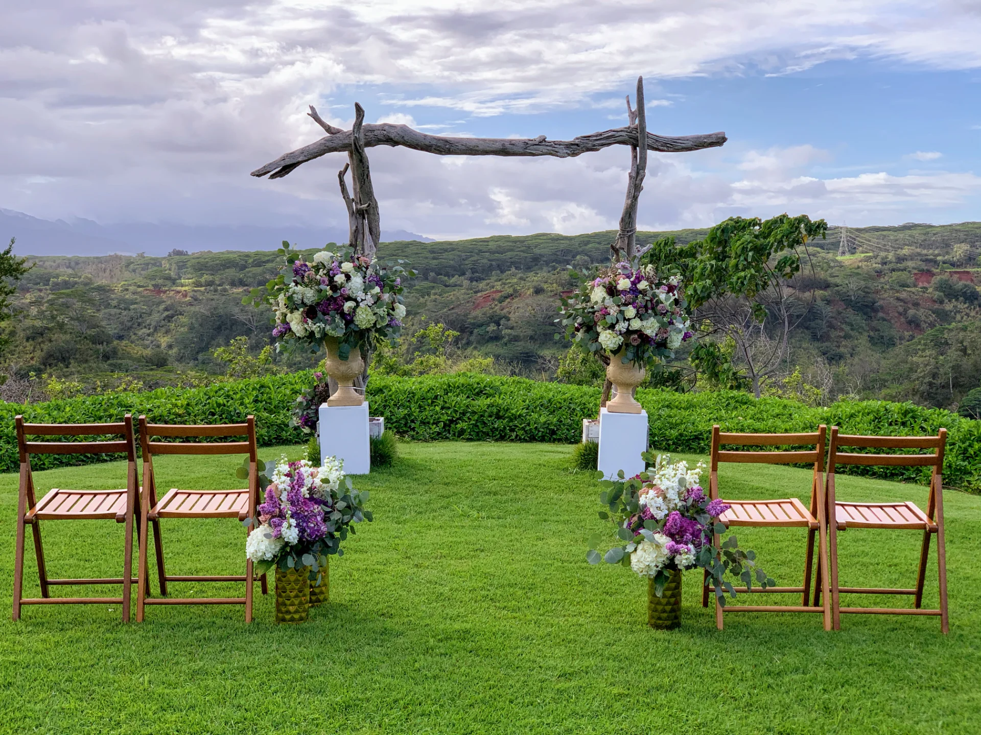 Luxury wedding ceremony setup in Oʻahu with rustic wooden arch and lush floral arrangements in purple and white hues.
