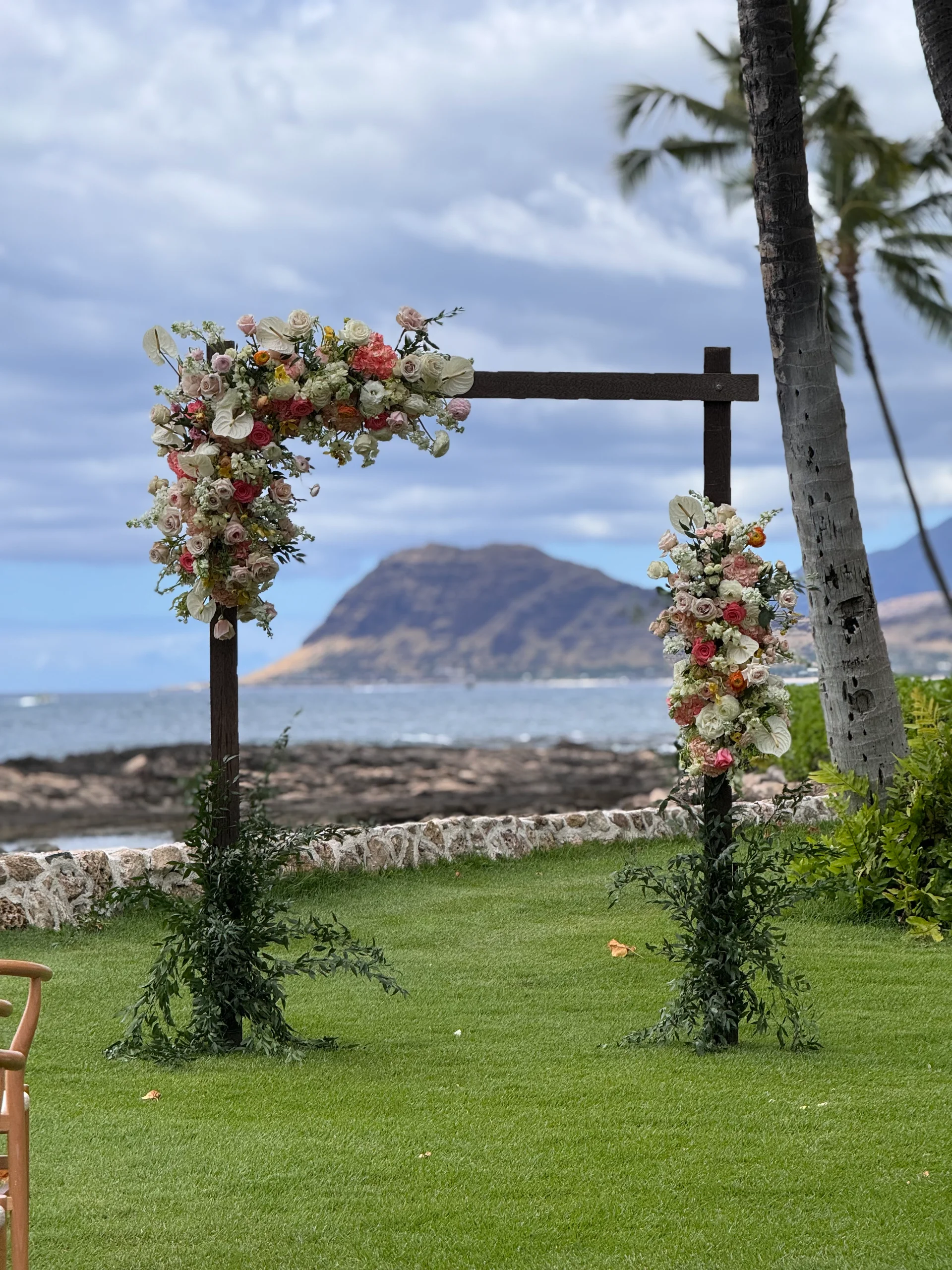 Floral arch with pink and white roses and greenery on a grassy lawn overlooking the ocean on Oʻahu.