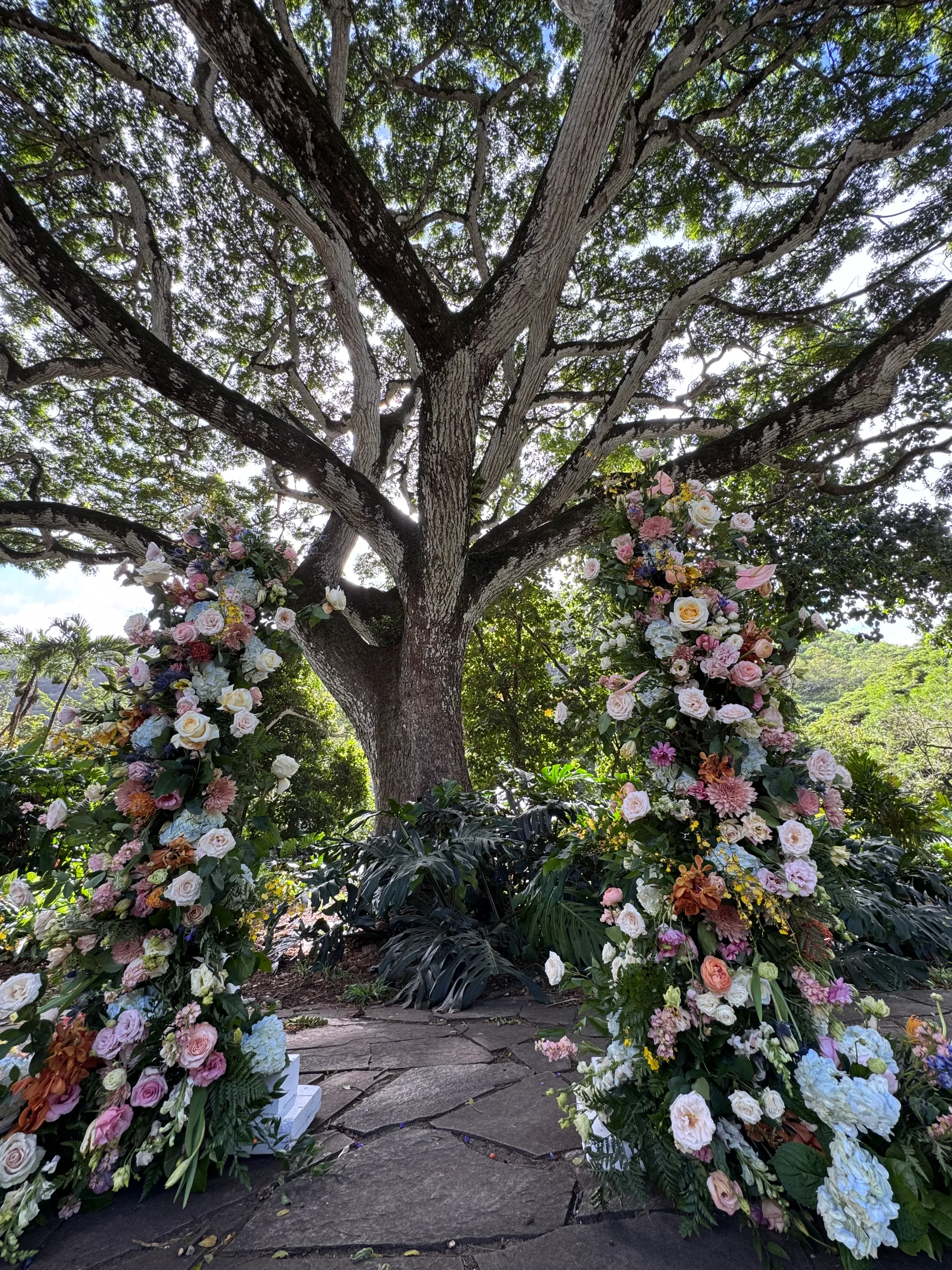 Lush floral arrangements with roses and hydrangeas under a large tree at an outdoor Hawaii wedding venue.