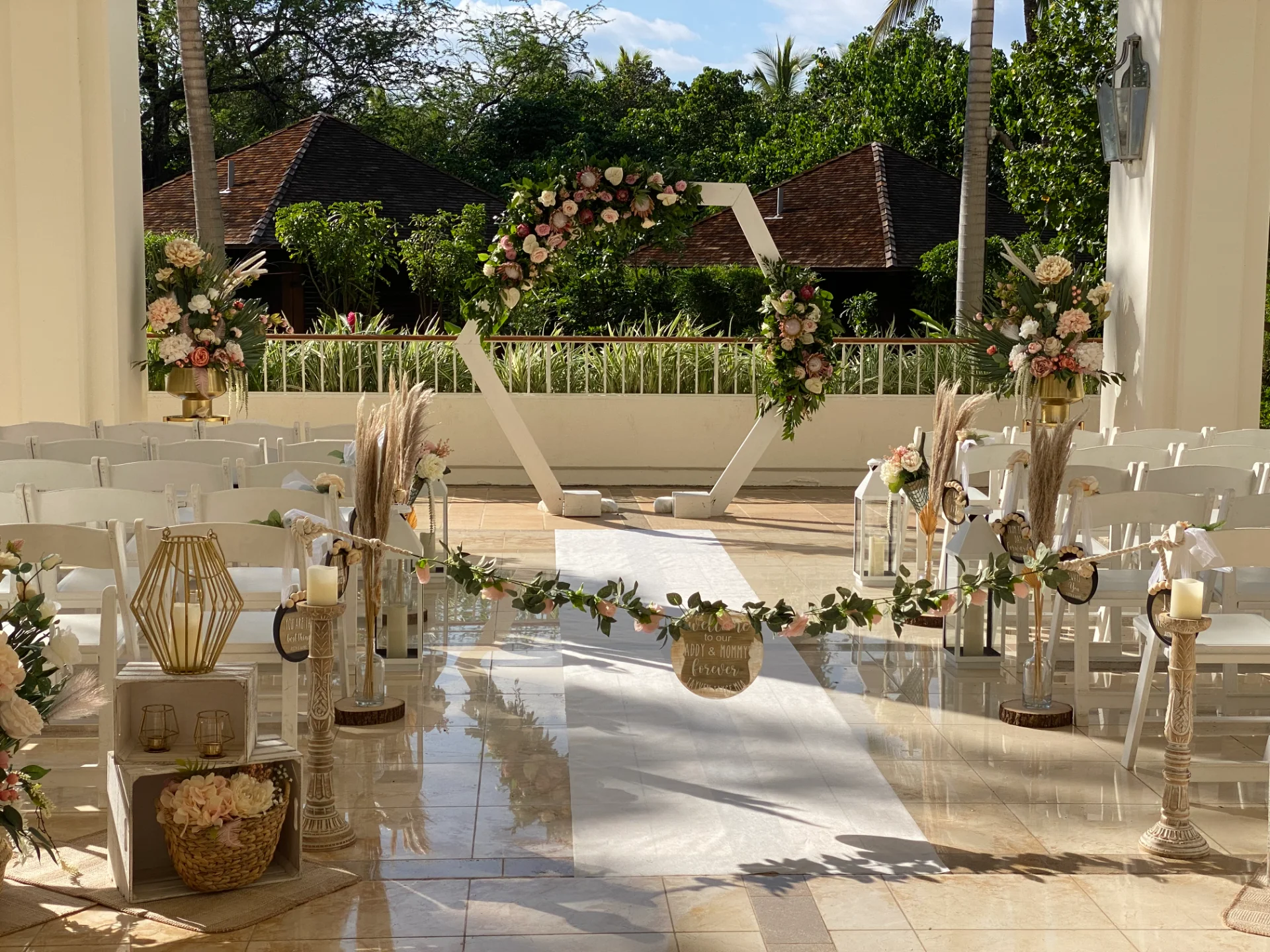 Outdoor wedding ceremony setup on Oʻahu with hexagonal floral arch featuring pink roses and greenery, white chairs, and pampas grass decor.