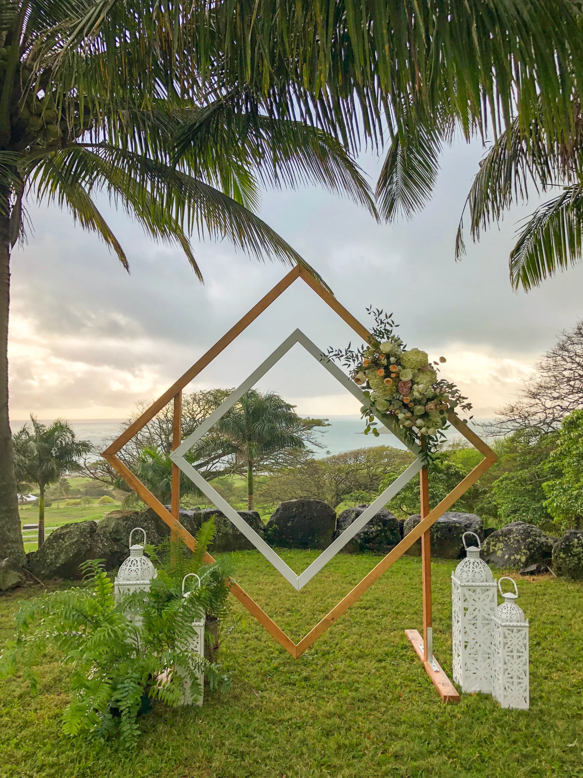 Geometric wedding arch with tropical floral arrangement set against an ocean view under palm trees, Oʻahu.