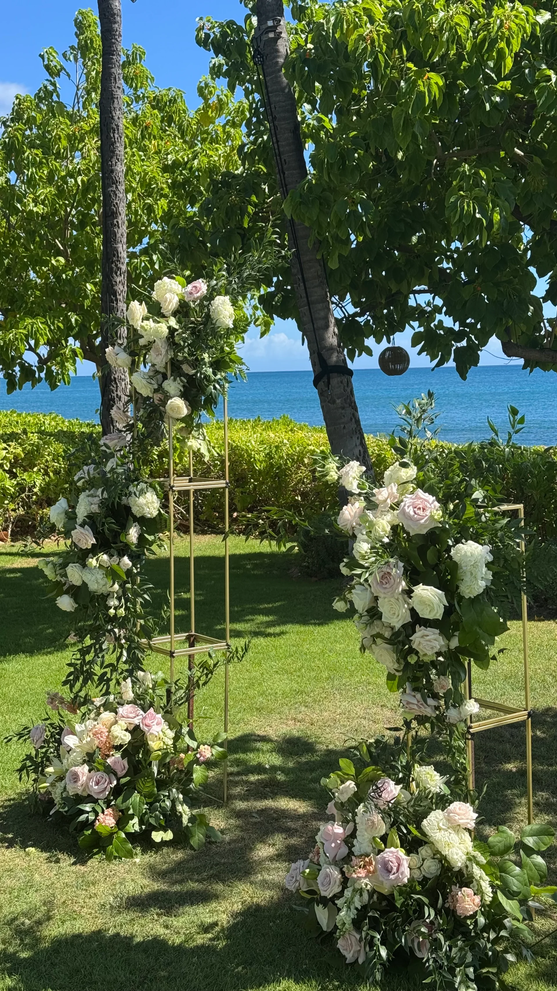 Elegant wedding floral display on Oʻahu with white and blush roses, greenery, and gold arches overlooking the ocean.