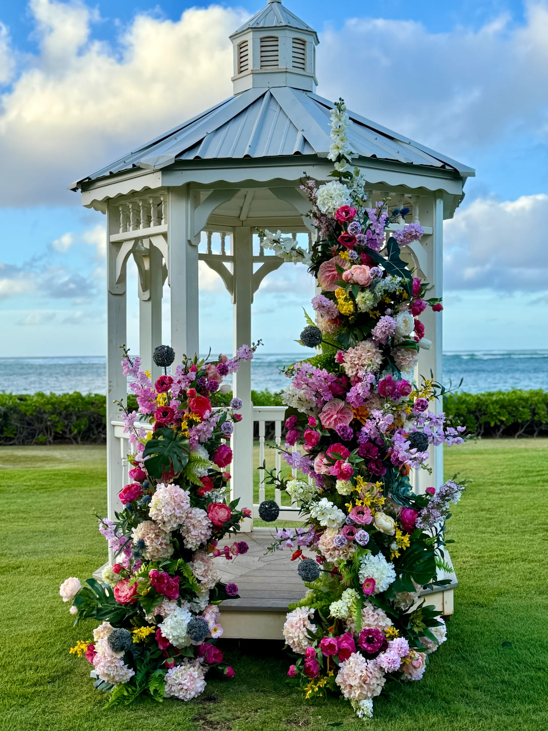 Elegant gazebo adorned with vibrant pink, white, and purple wedding florals on a lush lawn by the ocean in Oʻahu, Hawaii.