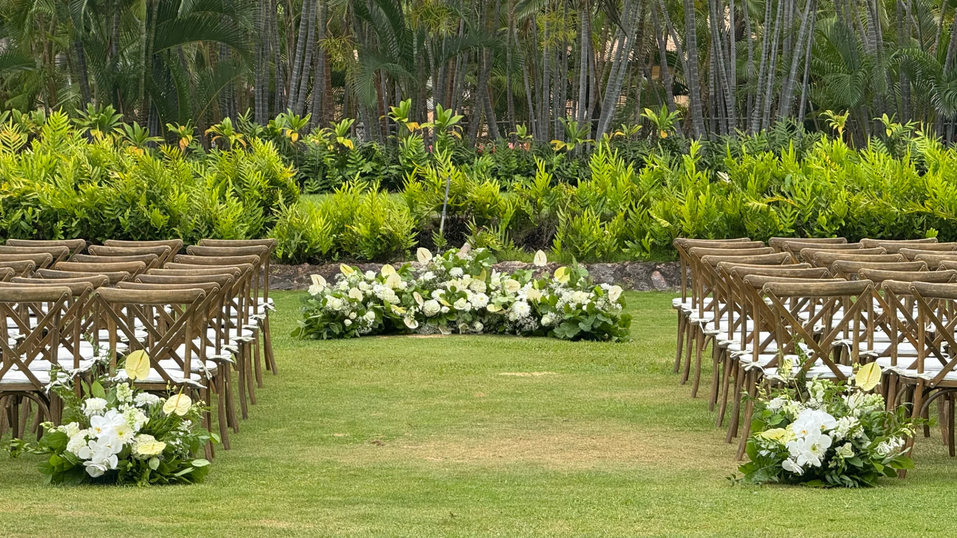 Elegant outdoor wedding setup with white florals and greenery lining a grass aisle on Oʻahu