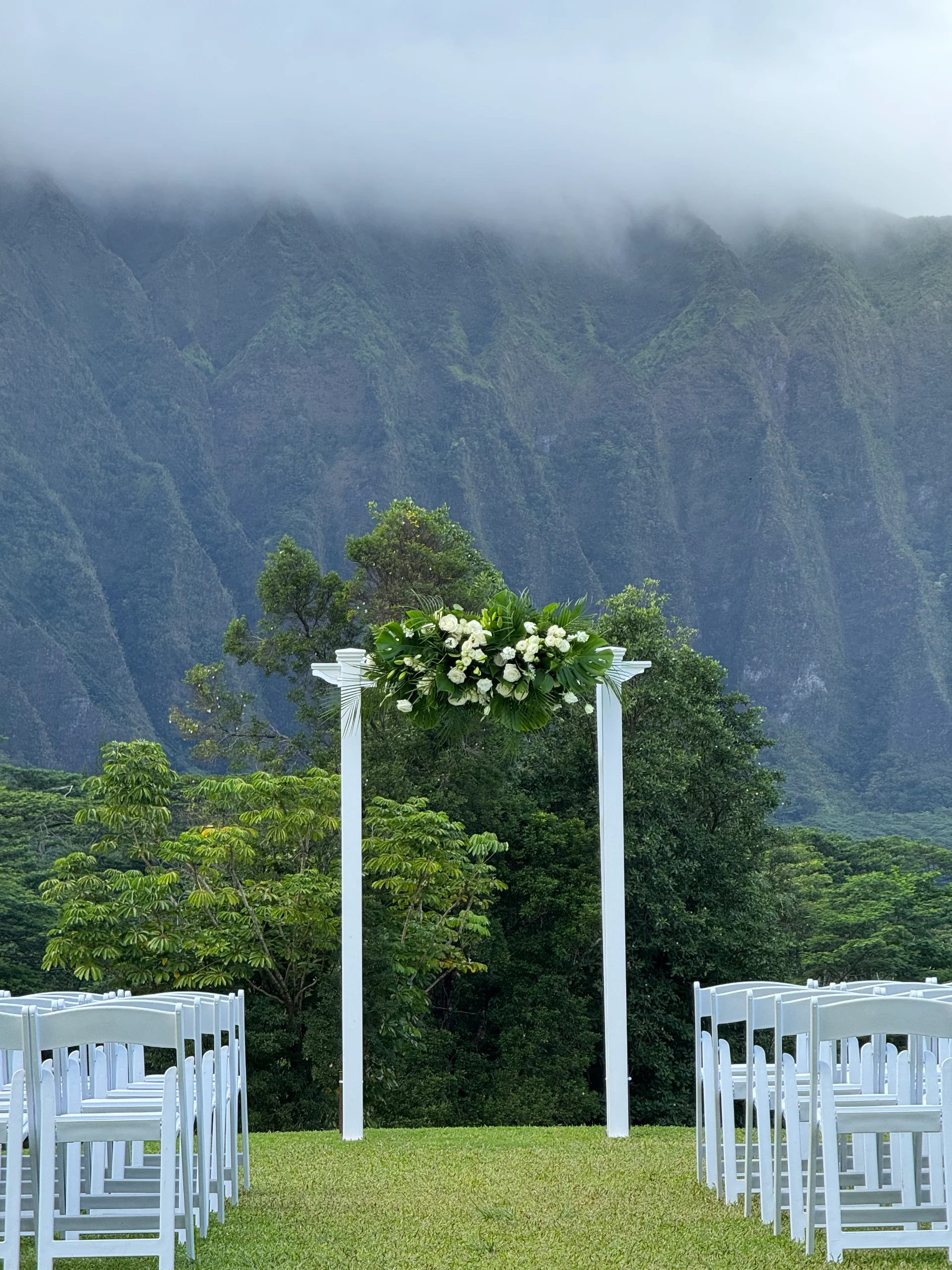Elegant outdoor wedding venue in Oʻahu with white floral arch and mountain backdrop.