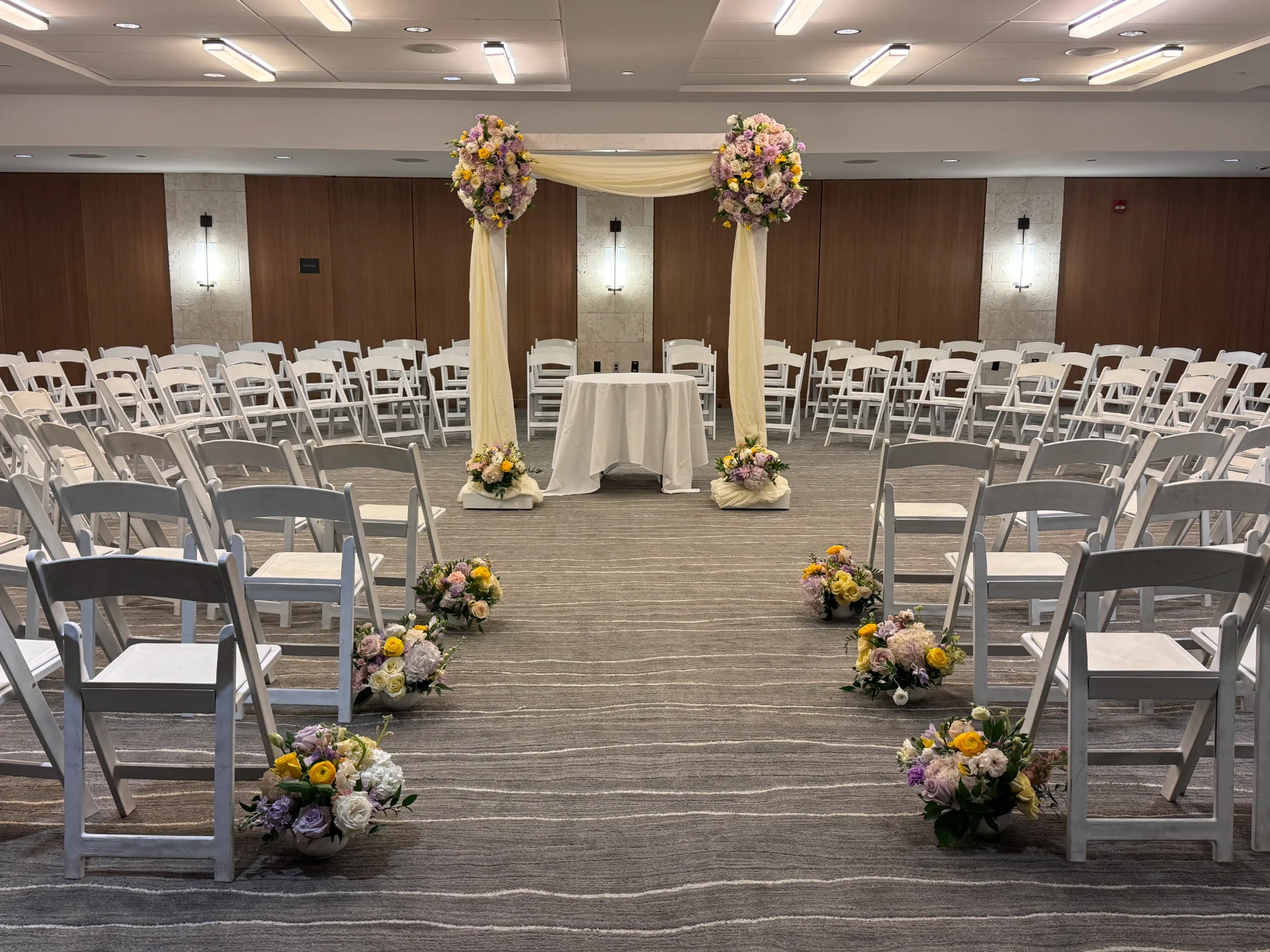 Indoor wedding ceremony setup with white chairs, floral aisle decor, and a flower-covered arch in soft pastels on Oʻahu