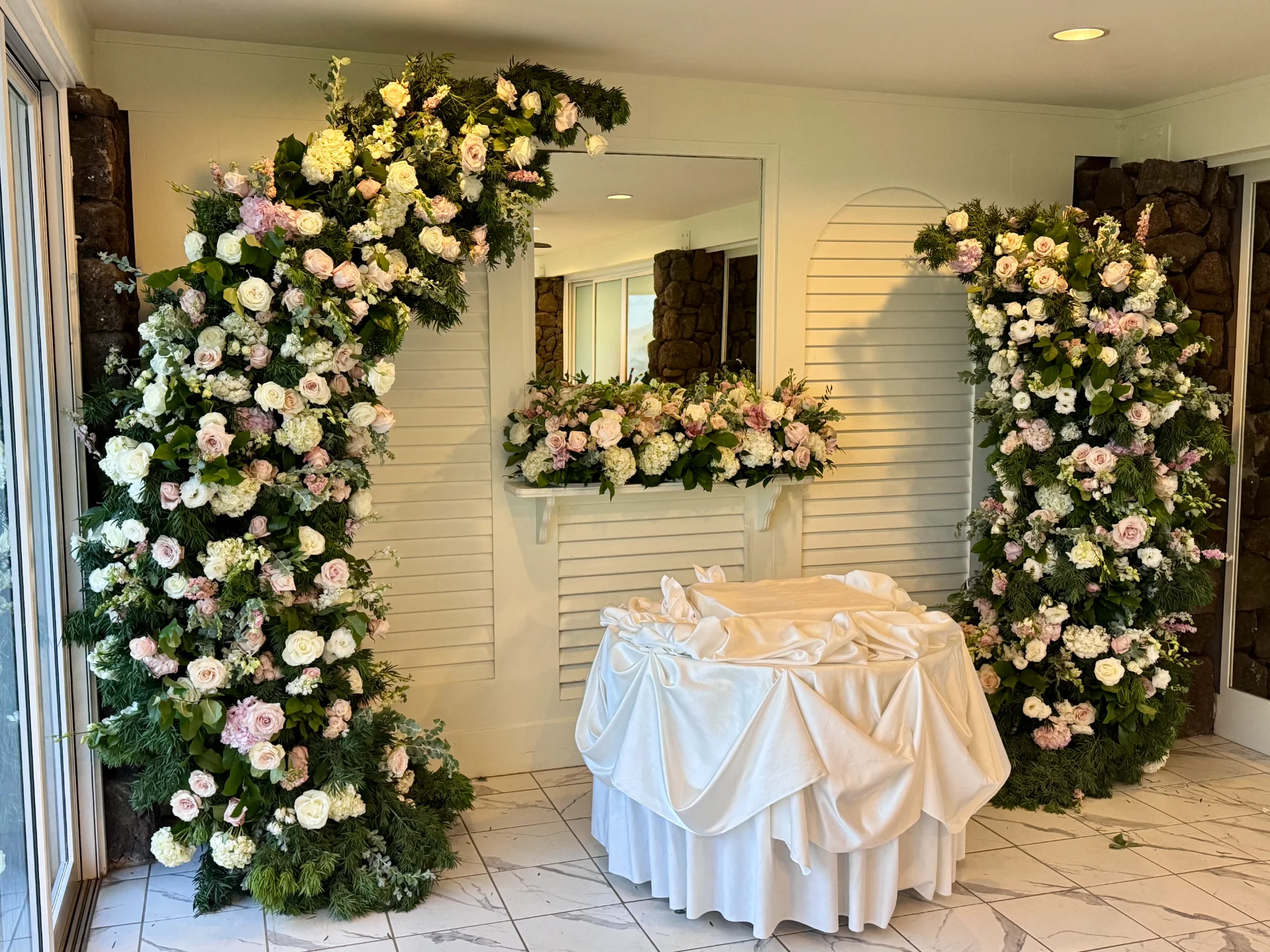 Elegant wedding floral arch with white and pink roses and greenery set indoors on Oʻahu, featuring a draped table and mirror decor