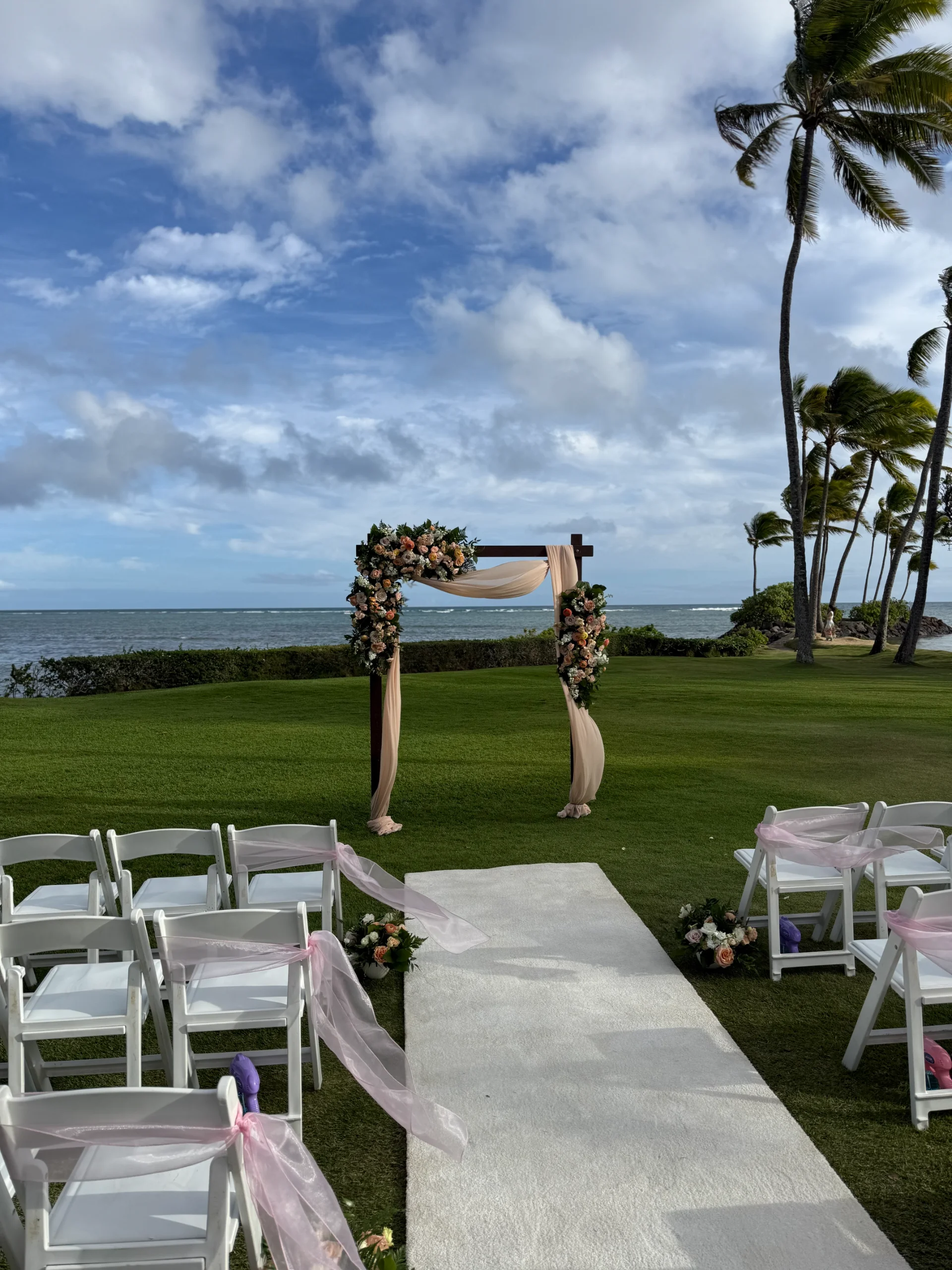 Beachfront wedding arch adorned with peach and white flowers, surrounded by palm trees and overlooking the ocean on Oʻahu.