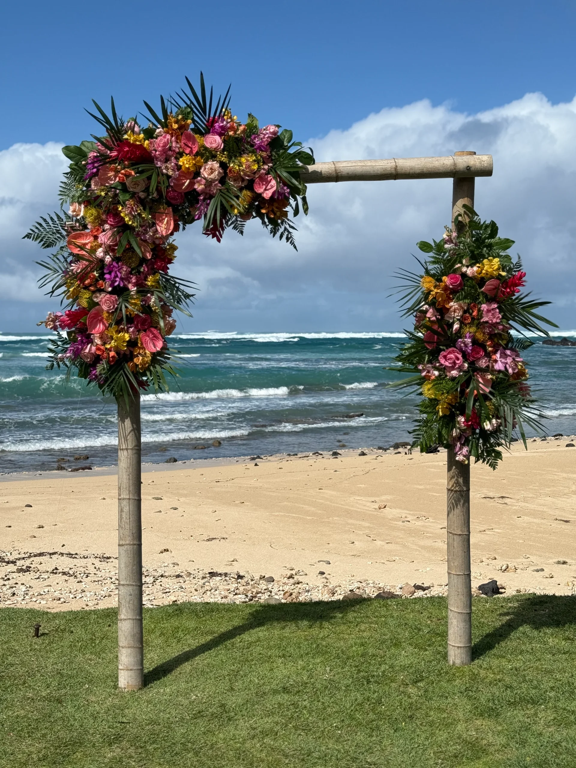 Tropical wedding arch adorned with vibrant pink, orange, and green florals on a beach in Oʻahu, Hawaii.