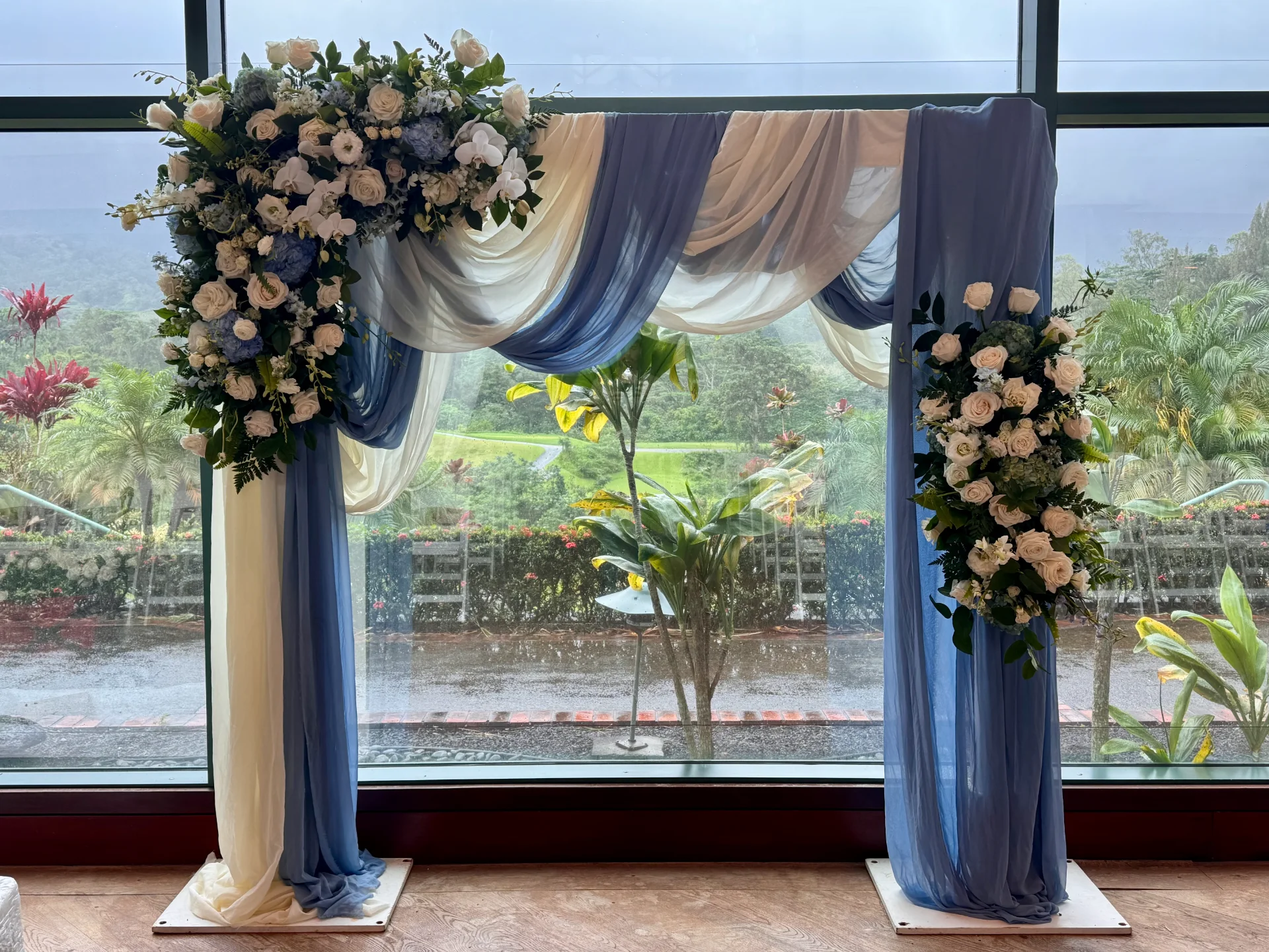 Elegant floral wedding arch with white and blue roses and draped fabric in front of a large window overlooking the lush Oʻahu landscape.