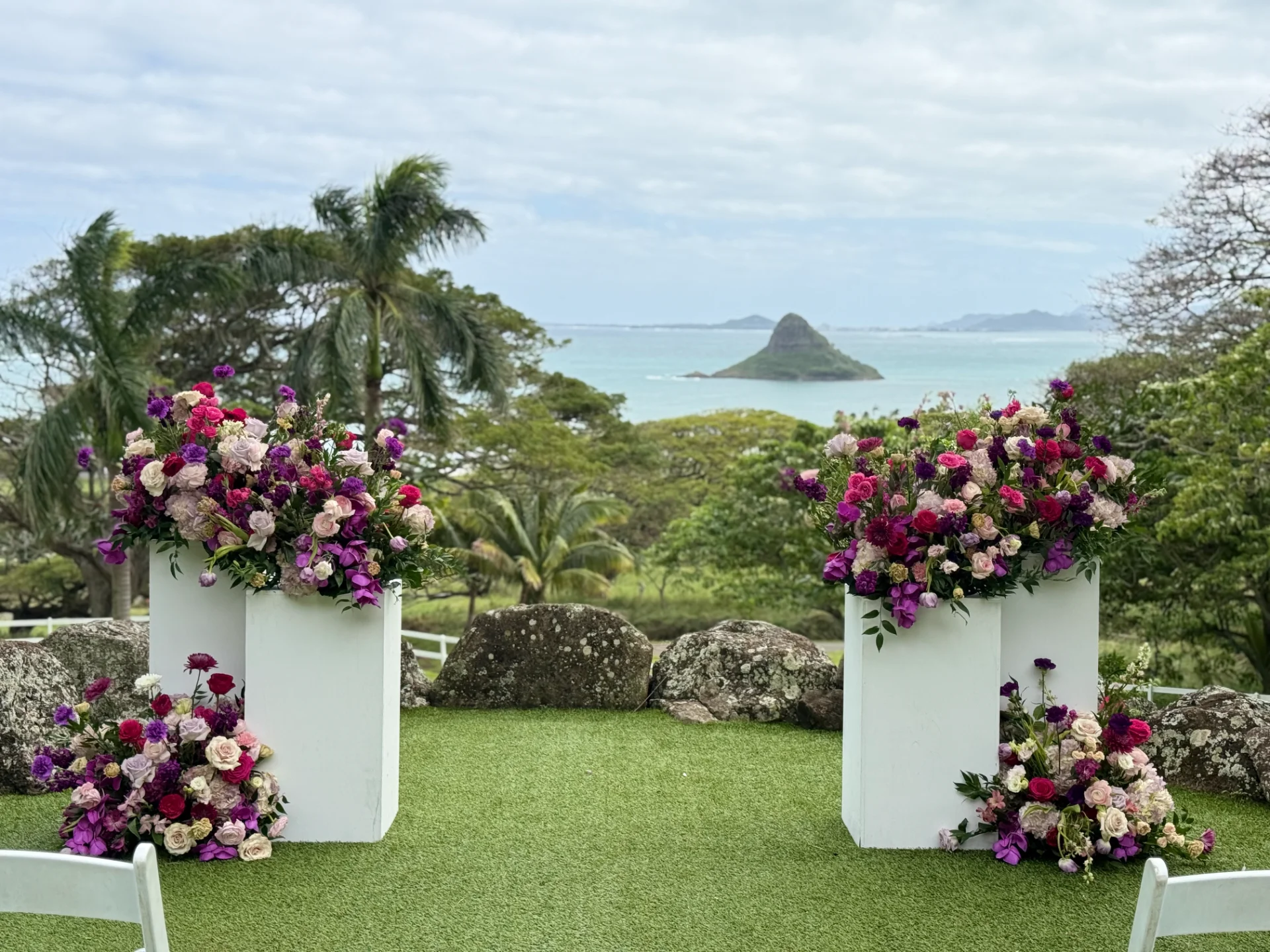 Elegant wedding floral arrangement with pink and purple roses on white pedestals overlooking Oʻahu coastline.