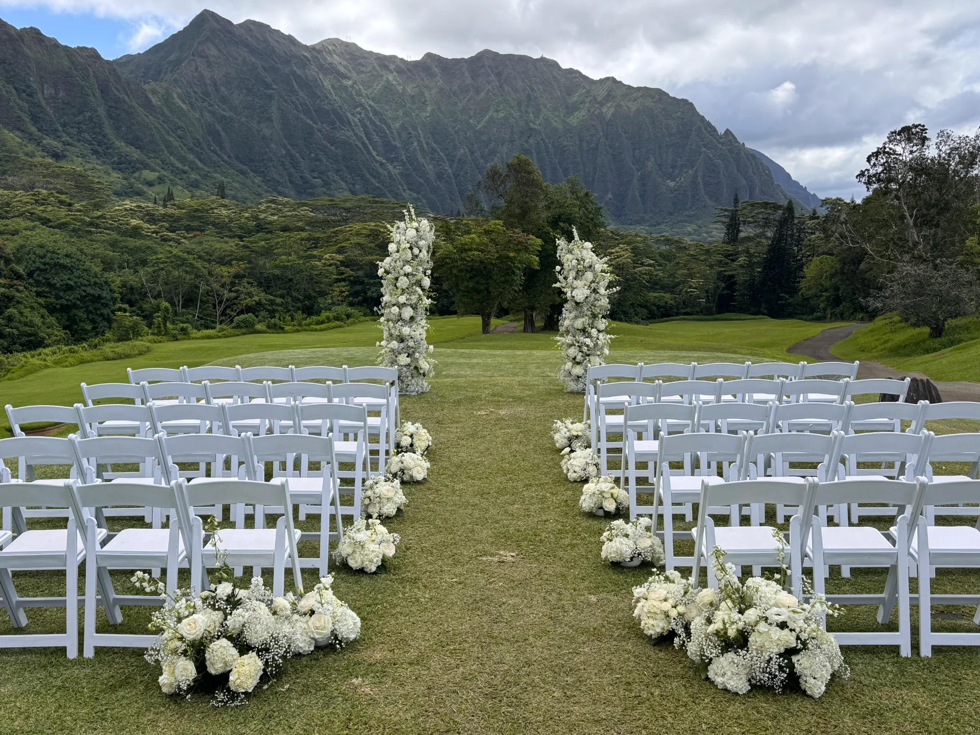 white chairs and floral aisle with white roses and baby's breath at outdoor wedding venue on Oʻahu with mountain backdrop