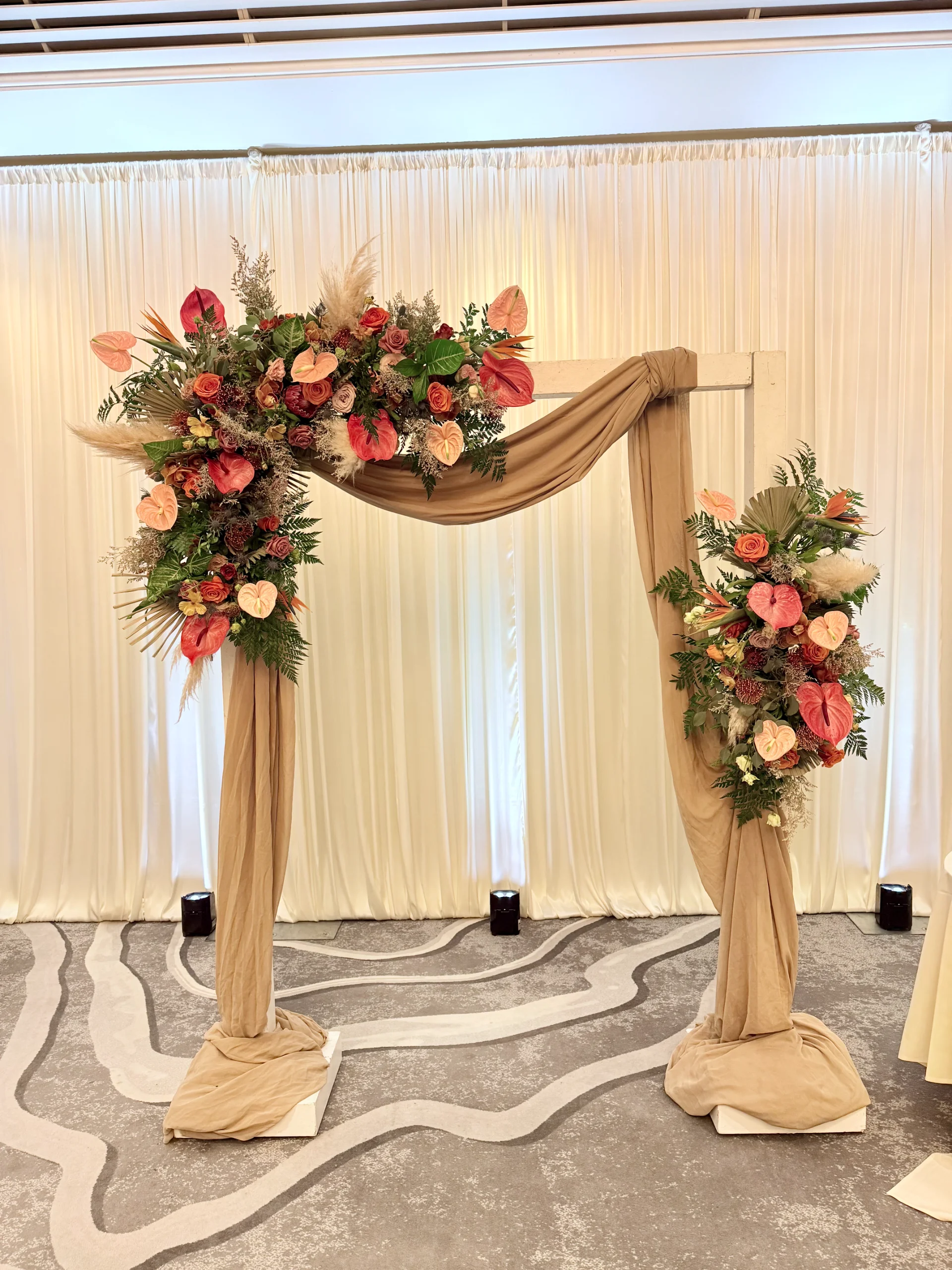 Elegant wedding arch with tropical flowers and beige drapery in Oʻahu, featuring anthuriums and roses against white curtains.