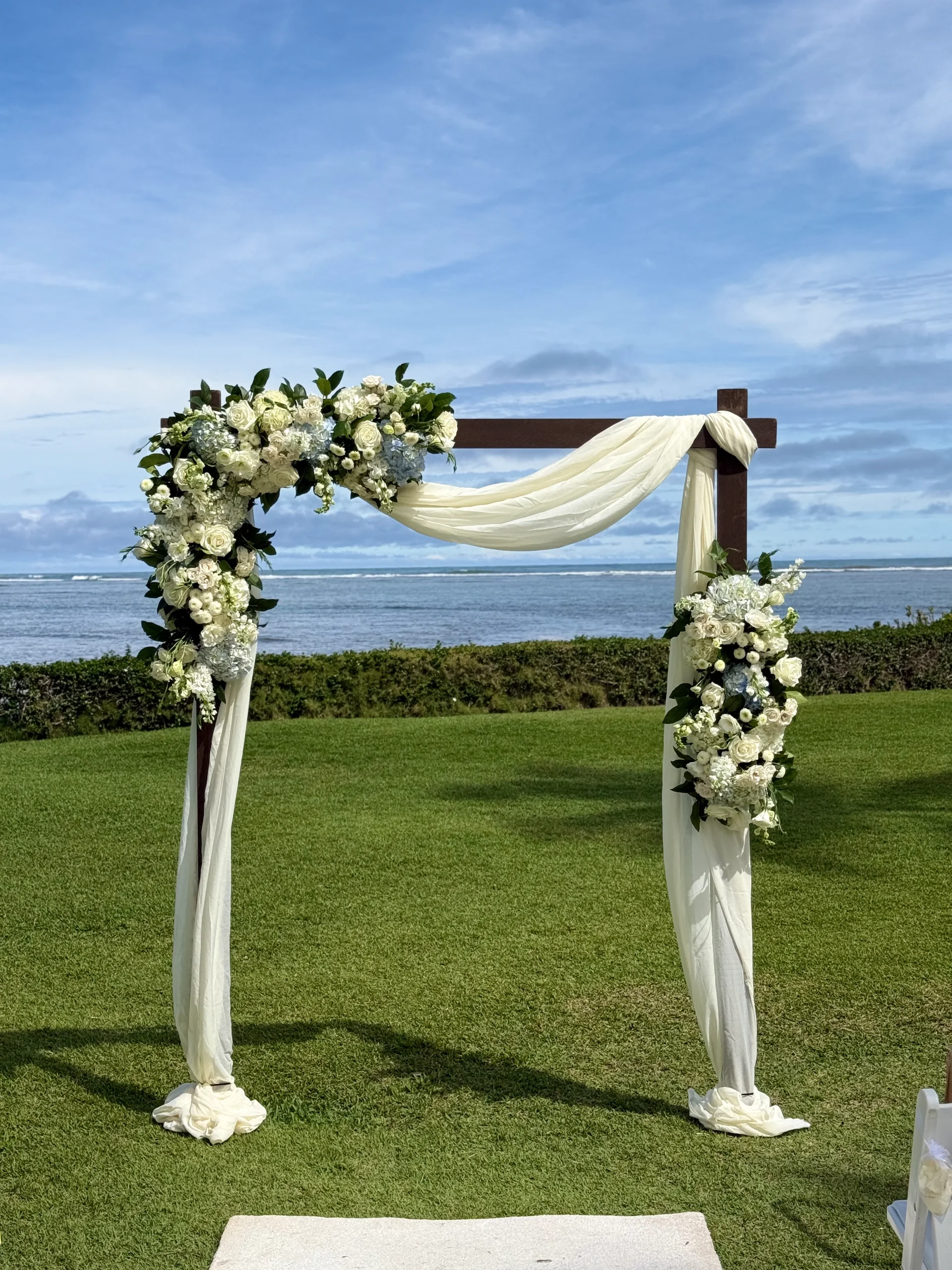 Beachside wedding arch in Oʻahu with white roses, hydrangeas, and draped fabric by the ocean.