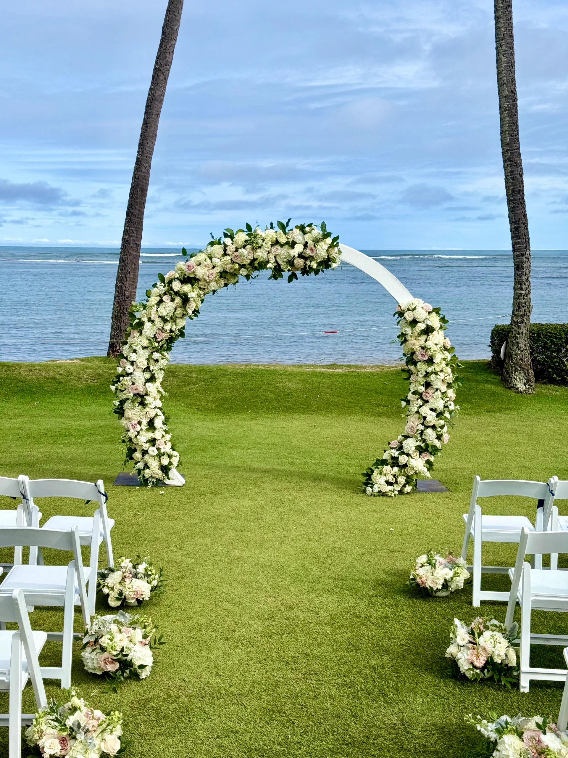 Elegant beachside wedding arch on Oʻahu adorned with white and blush roses, surrounded by white chairs and bouquets.
