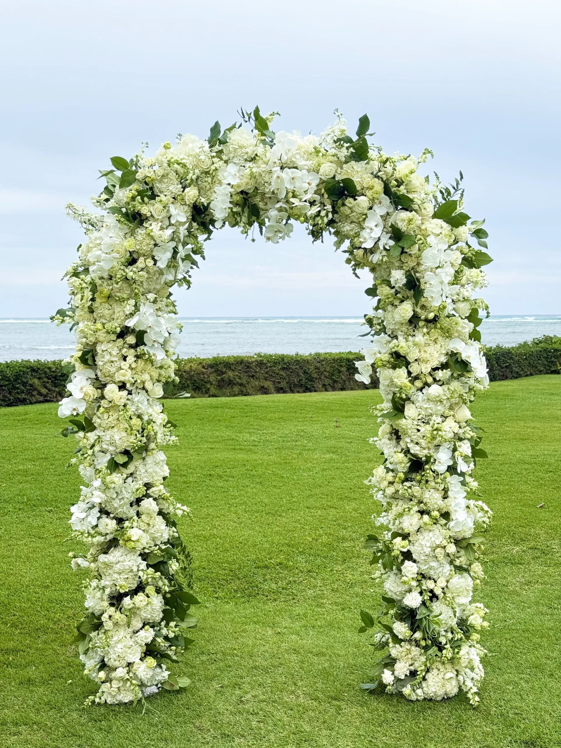Elegant floral arch with white roses and orchids on green lawn by the ocean in Oʻahu, Hawaii