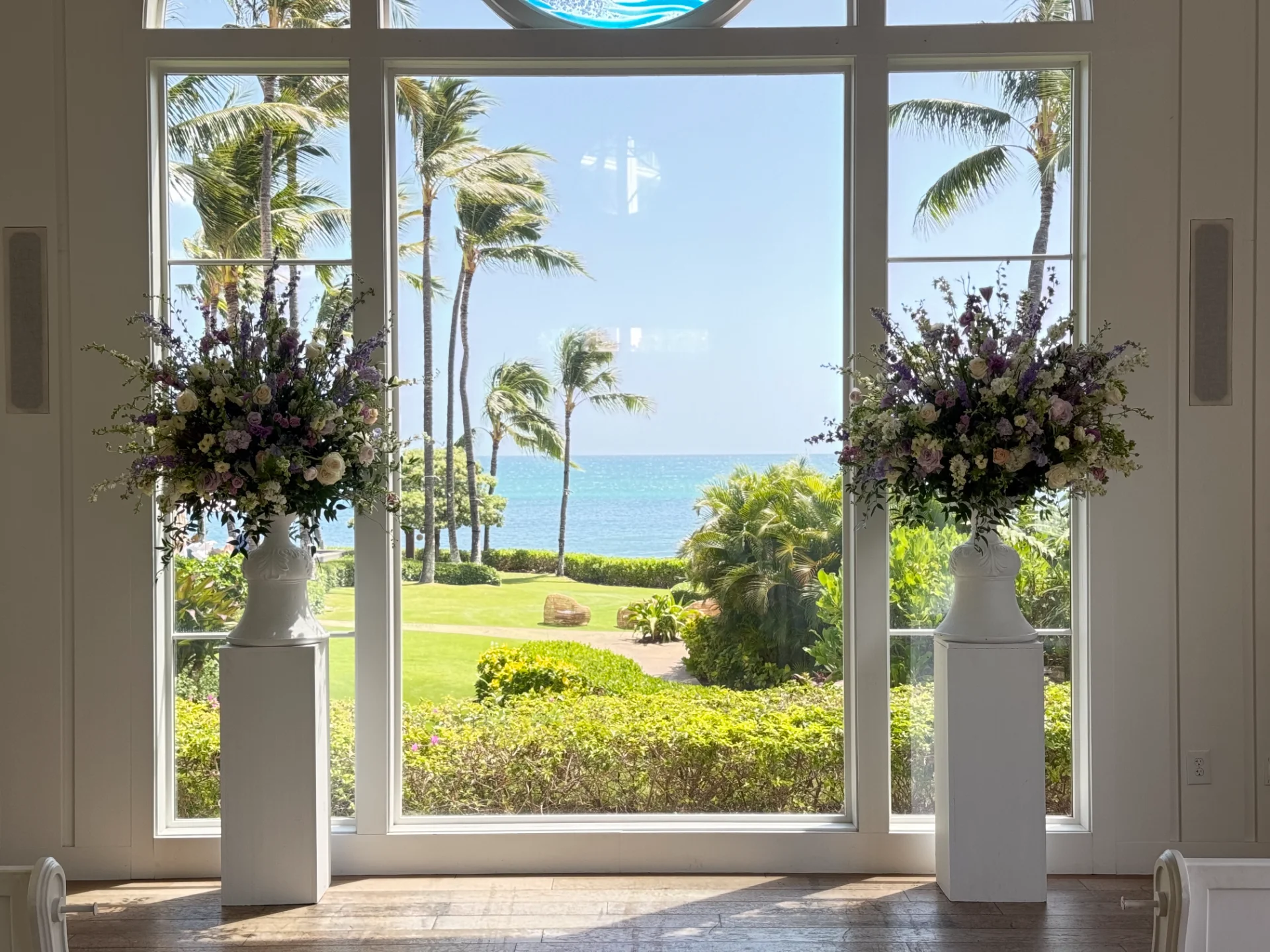 Elegant floral arrangements with purple and white flowers on pedestals in front of large window with ocean view in Oʻahu.