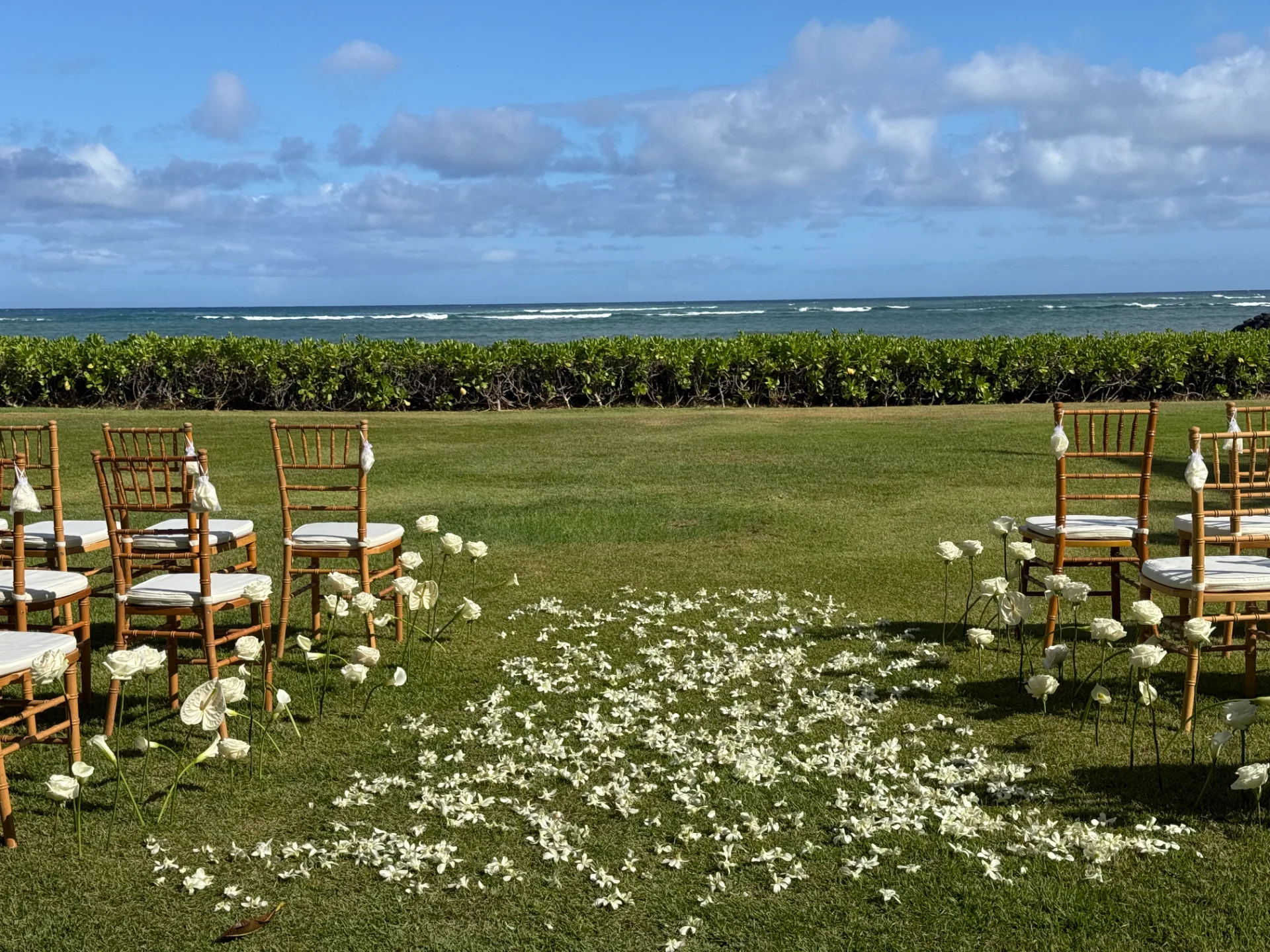 Oʻahu oceanfront wedding setup with white floral aisle, wooden chairs, and blue sky