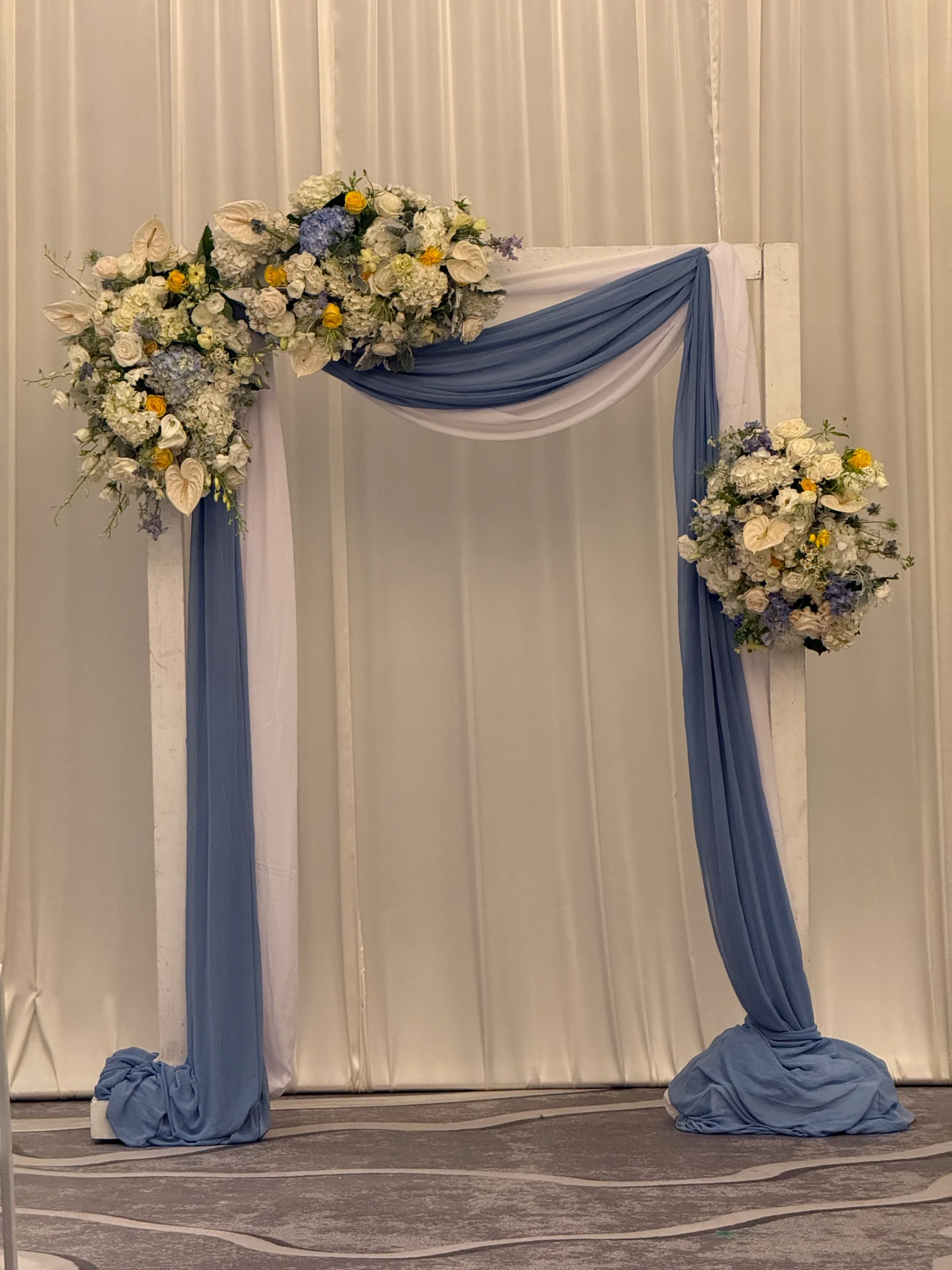 Elegant wedding arch with white and blue drapery adorned with white roses, anthuriums, yellow roses, and blue hydrangeas in Oʻahu.