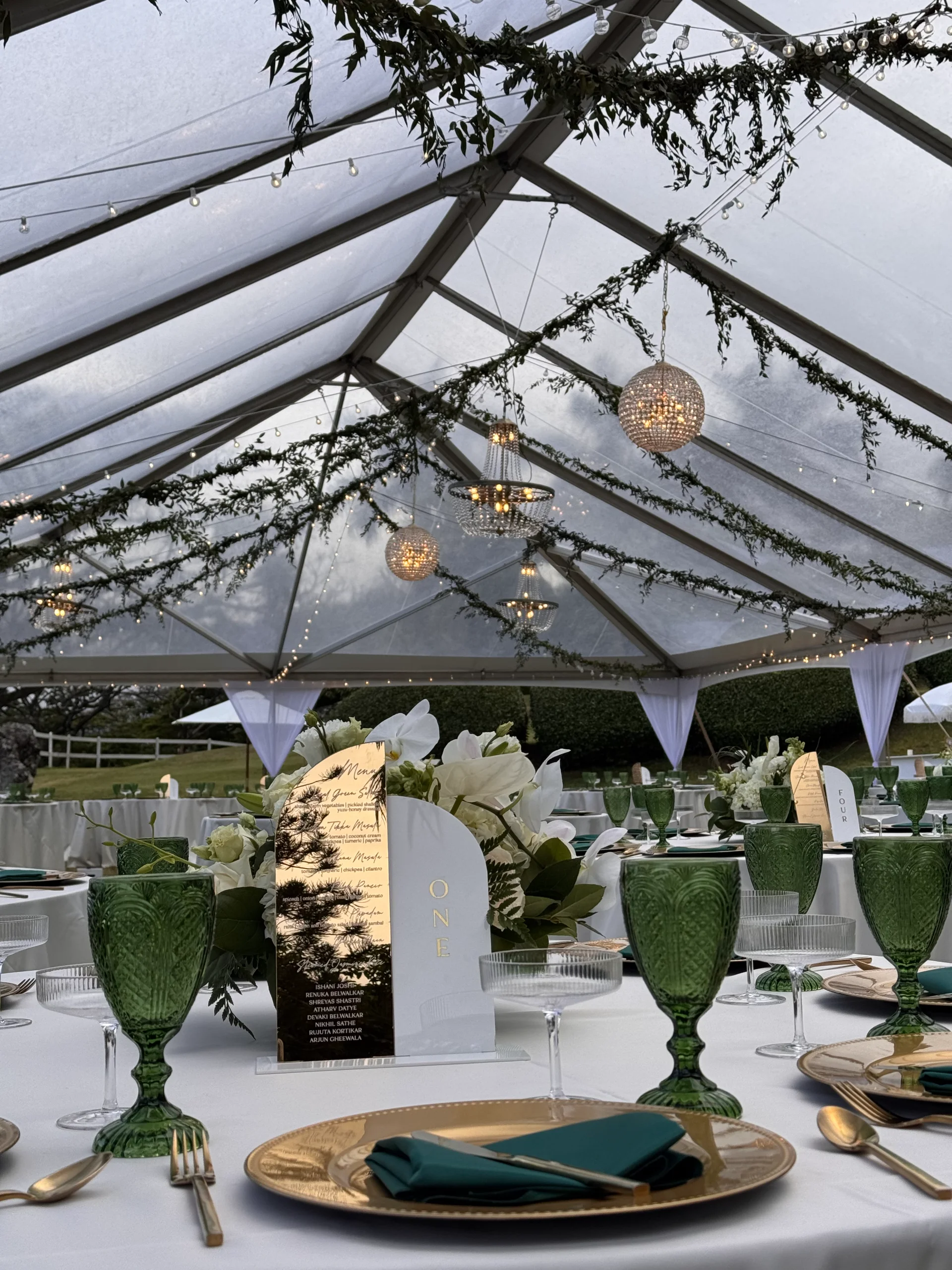 Elegant wedding reception under a transparent tent on Oʻahu with green glasses, gold chargers, and lush white floral centerpieces.