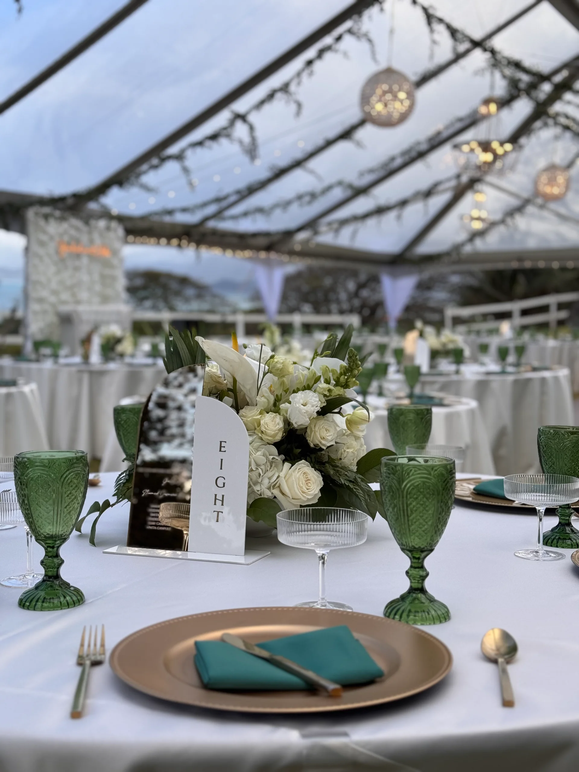 Elegant wedding reception setup on Oʻahu featuring white floral centerpieces, green glassware, teal accents, and string lighting.
