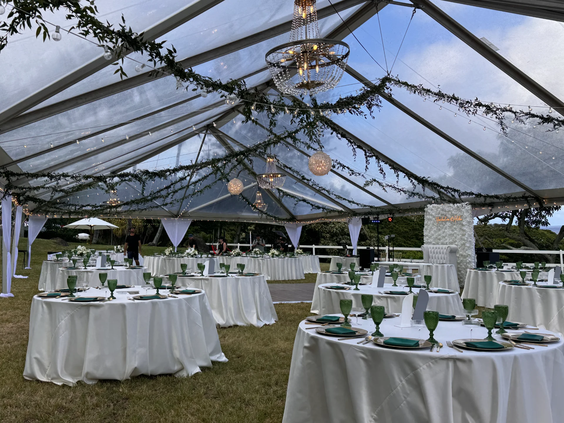 Elegant outdoor wedding reception under a transparent tent with chandeliers, green glassware, and white linens in Oʻahu