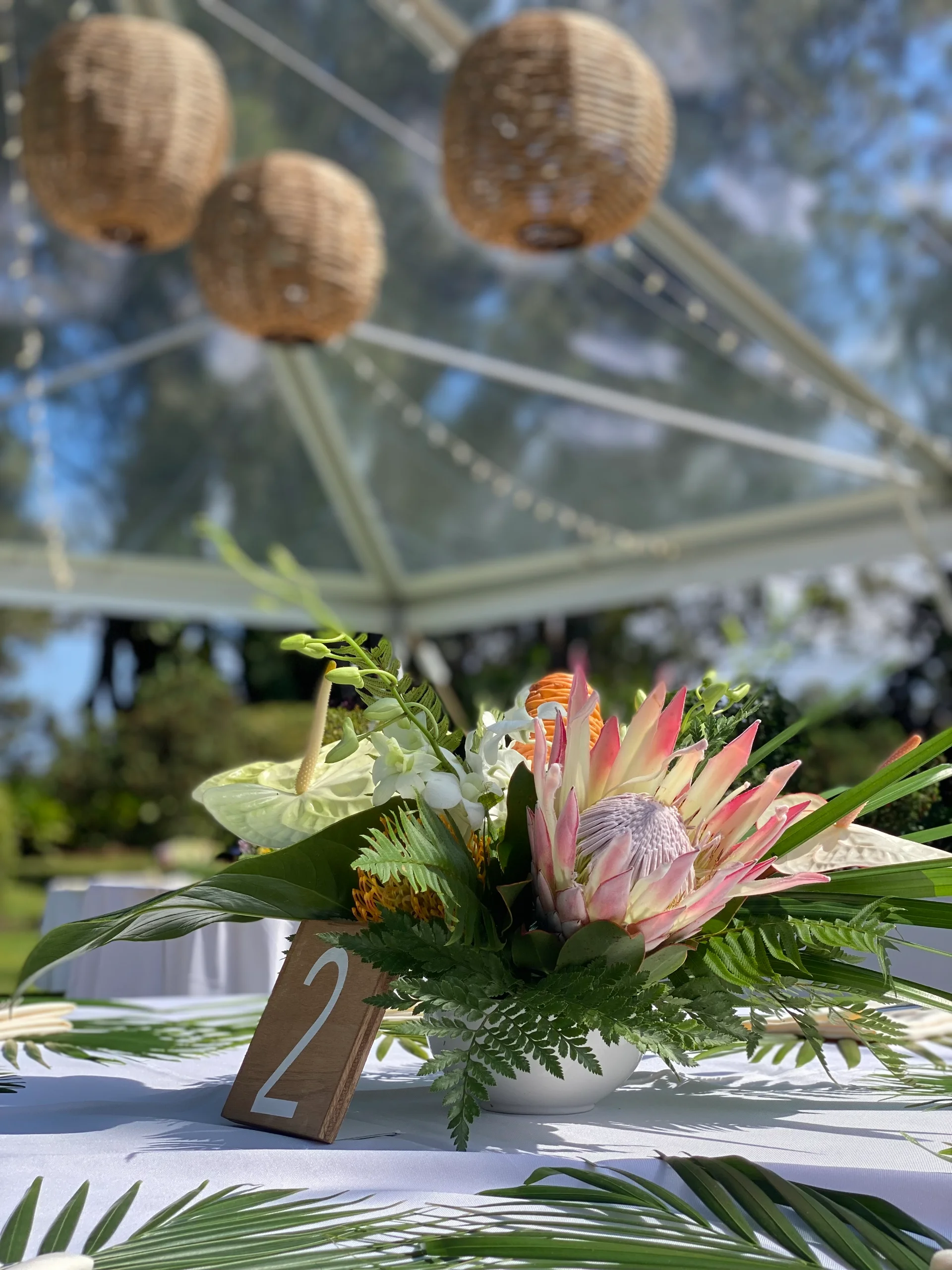 Hawaiian wedding centerpiece with protea, anthurium, and tropical greenery under a canopy with rattan lanterns on Oʻahu.