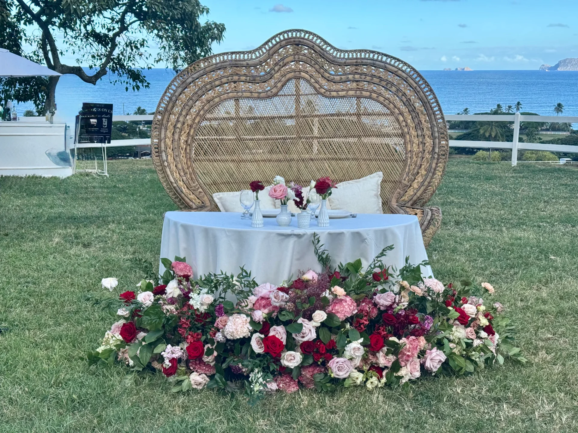 Ornate wicker loveseat and table adorned with red and pink roses, hydrangeas at outdoor wedding in Oʻahu, overlooking ocean.