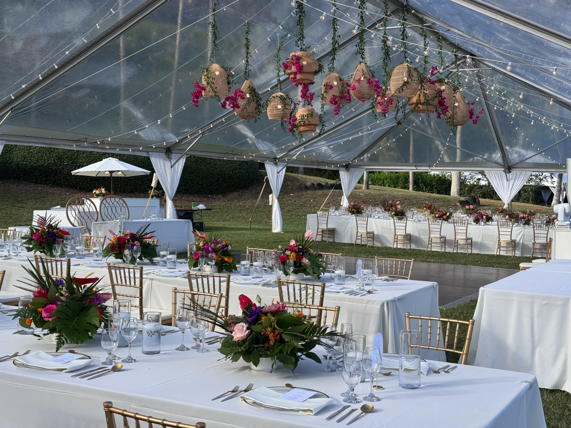 Luxury wedding reception with vibrant floral centerpieces under a clear tent on Oʻahu, featuring wicker lanterns and white settings.