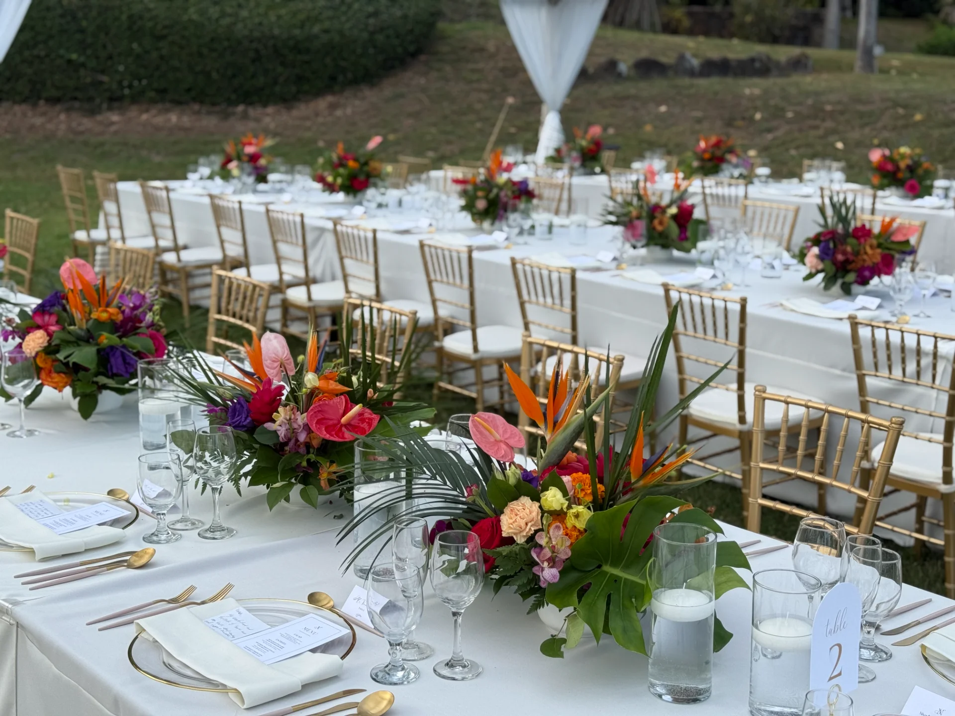 Elegant table setting with vibrant tropical floral centerpieces and gold chairs at an outdoor Oʻahu wedding reception