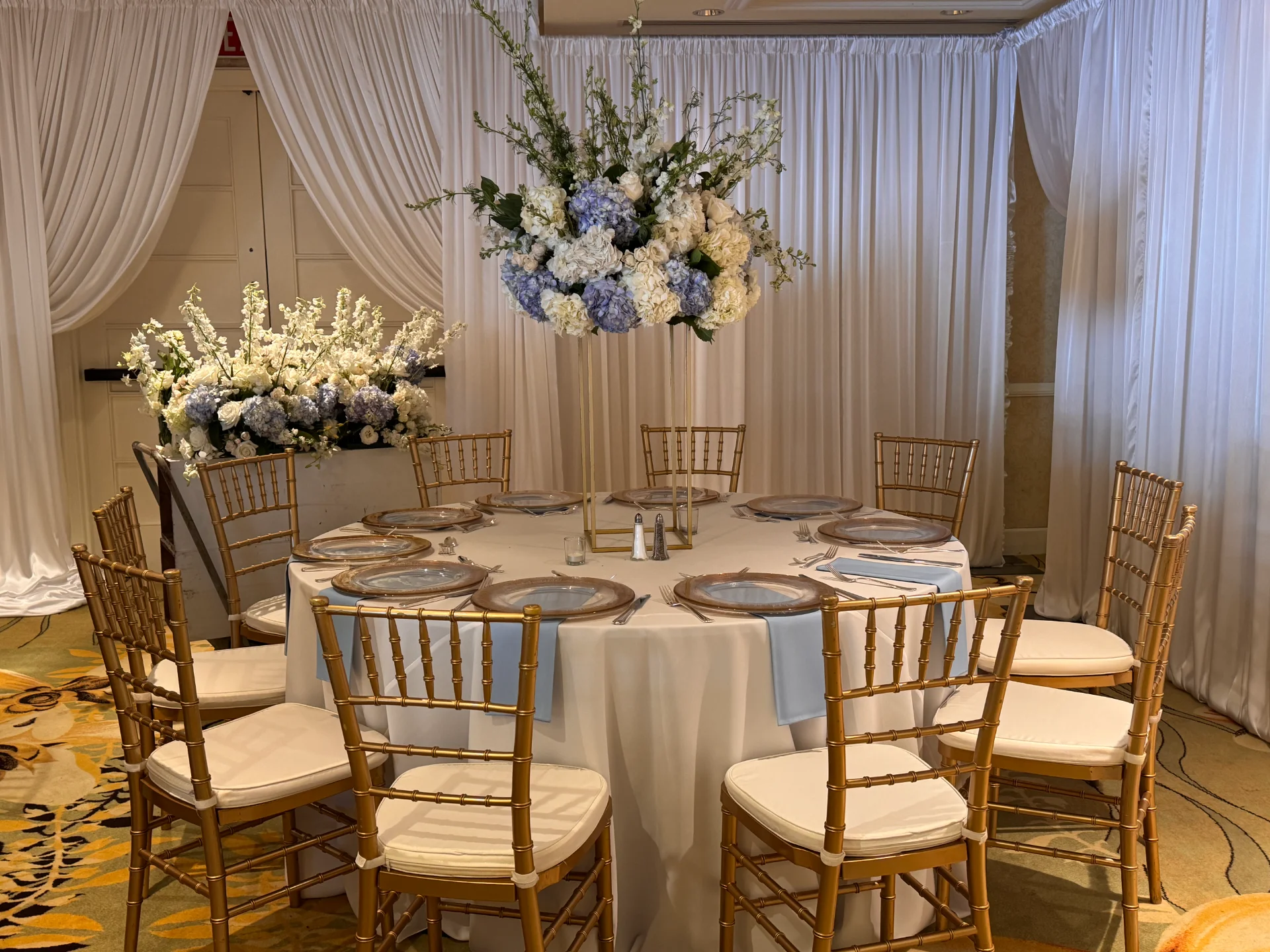 Elegant wedding reception table with gold chairs, white and pale blue floral centerpiece, draped white curtains in Oʻahu.