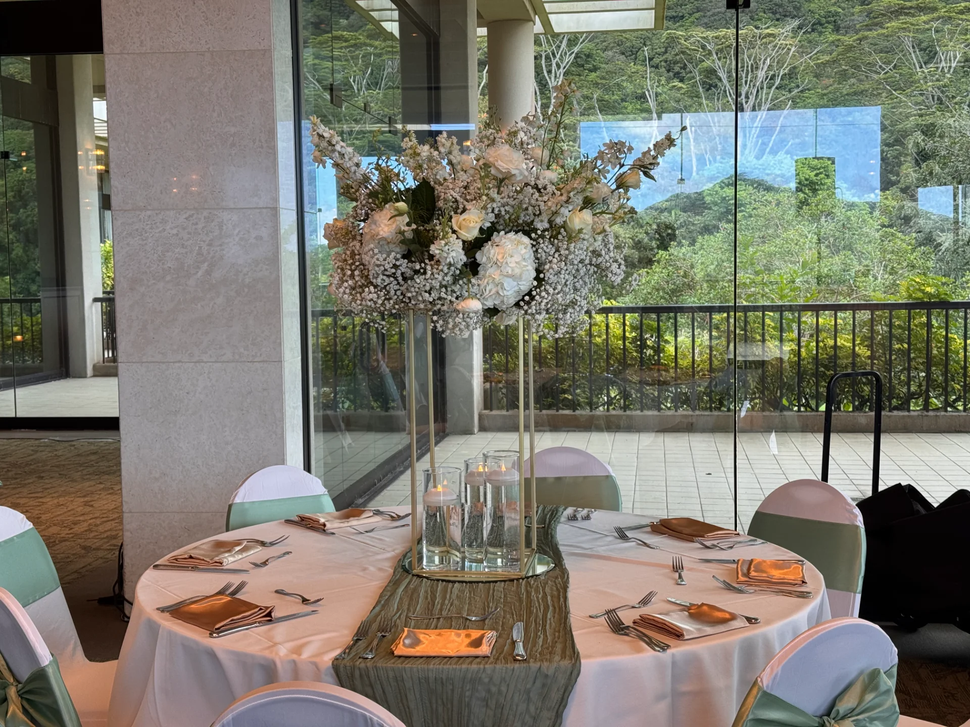 Elegant wedding reception table with tall floral centerpiece featuring white roses, hydrangeas, and baby's breath, set in Oʻahu venue with lush greenery view.
