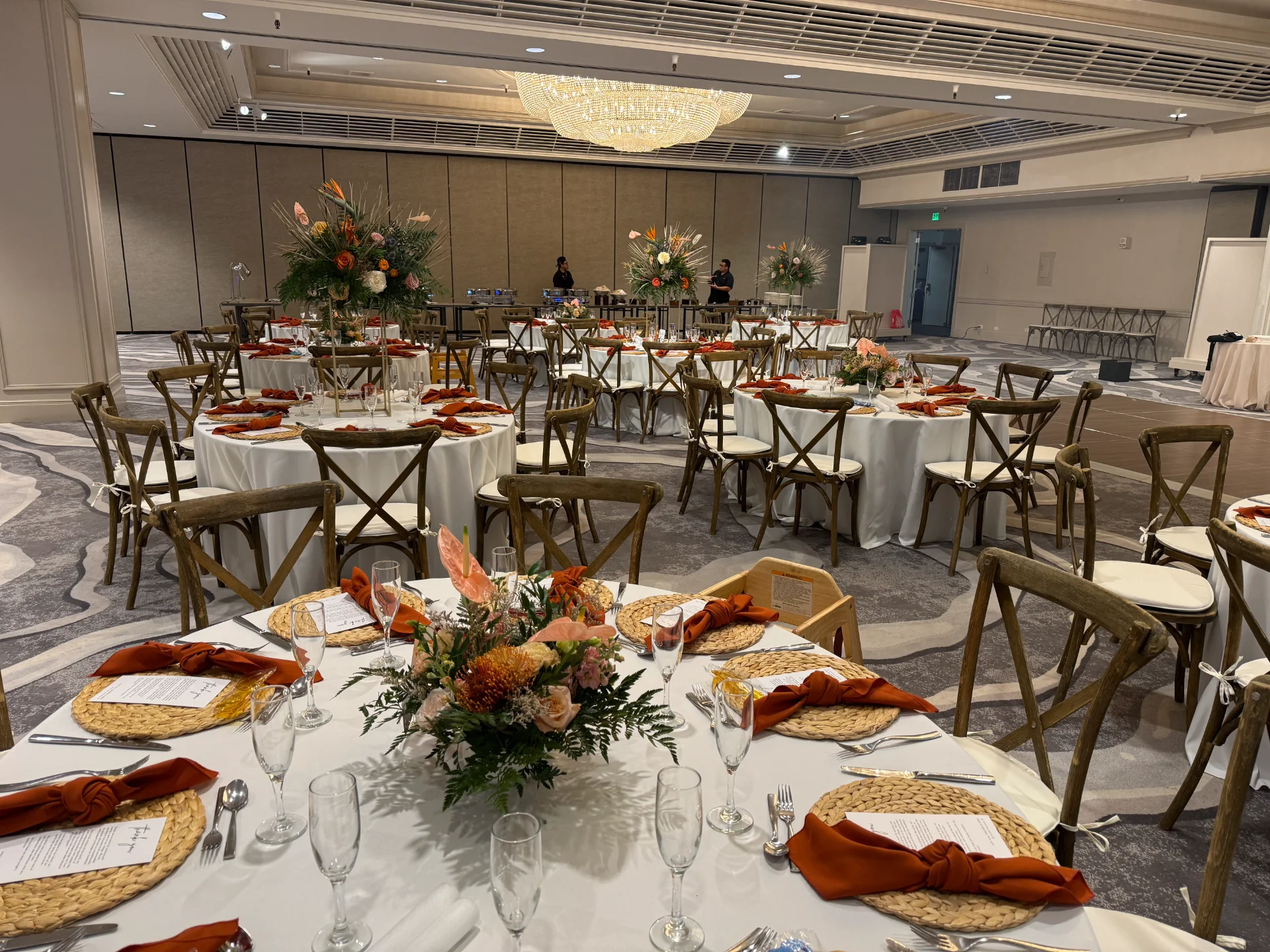 Elegant wedding reception setup in Oʻahu with round tables, vibrant floral centerpieces, and orange napkins under a chandelier.
