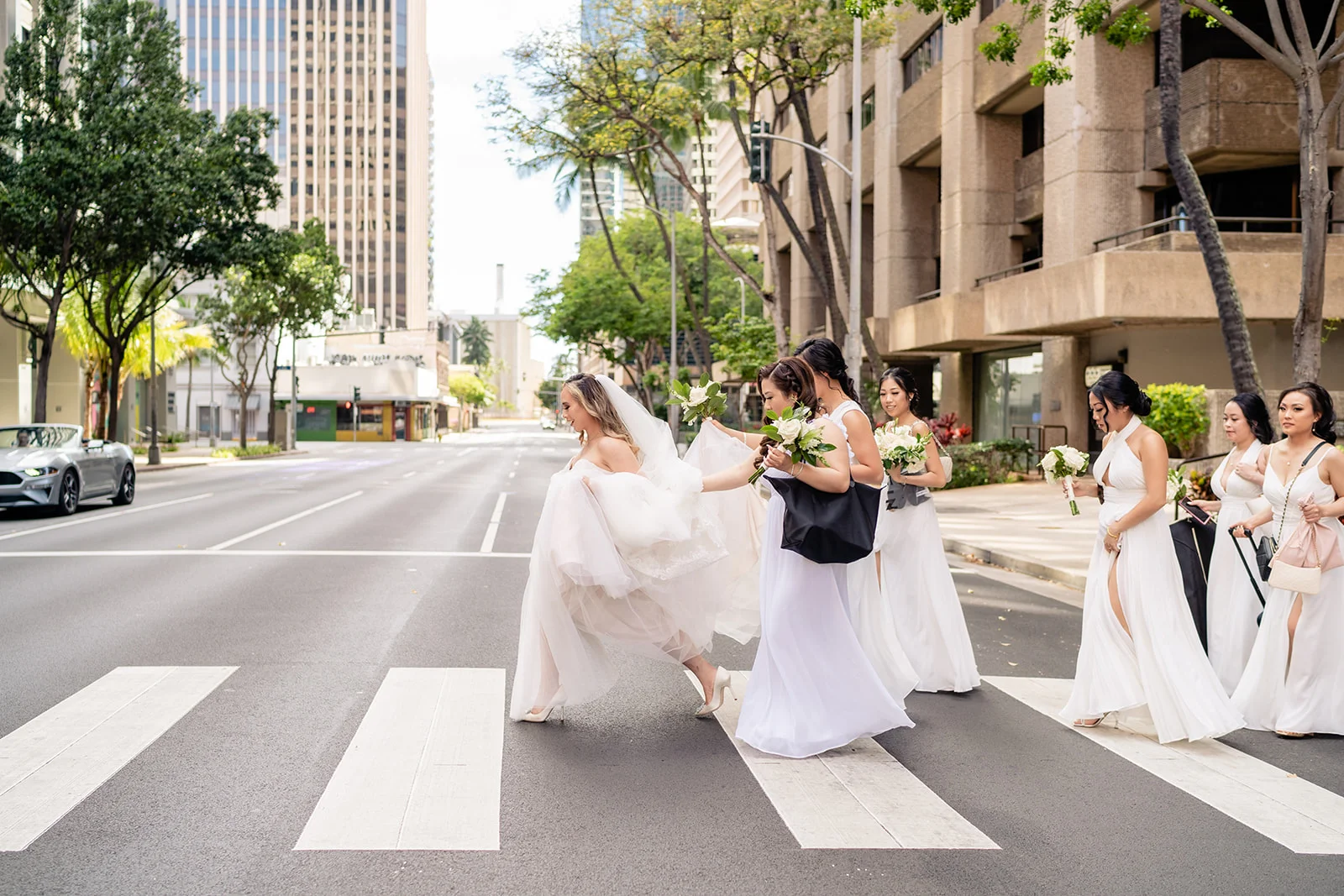 Bridal party in white dresses crossing a city street with bouquets at Cafe Julia on Oʻahu, Hawaii.