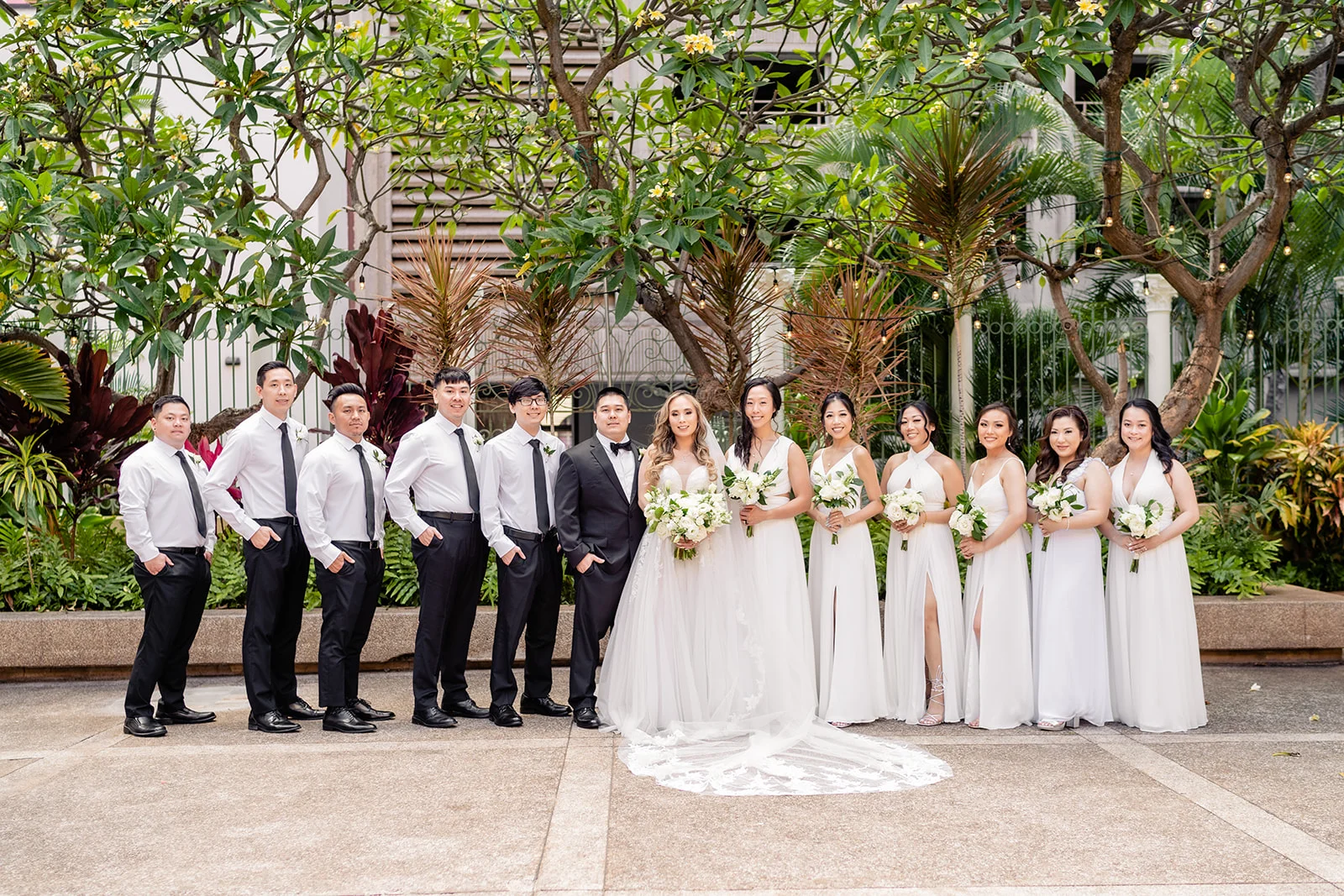 Bridal party at Cafe Julia in Oʻahu surrounded by lush greenery, wearing formal attire with white and green floral bouquets