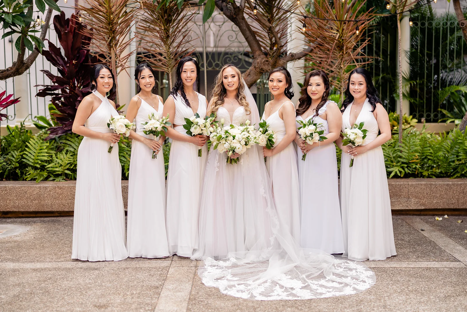 Bride and bridesmaids in white dresses holding white and green bouquets at Cafe Julia, Oʻahu wedding setting.