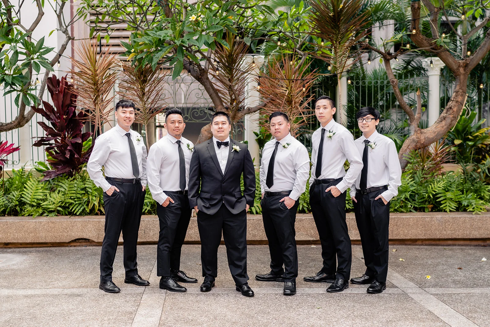 Groomsmen in black suits and white shirts stand in an outdoor garden setting at Cafe Julia in Oʻahu.