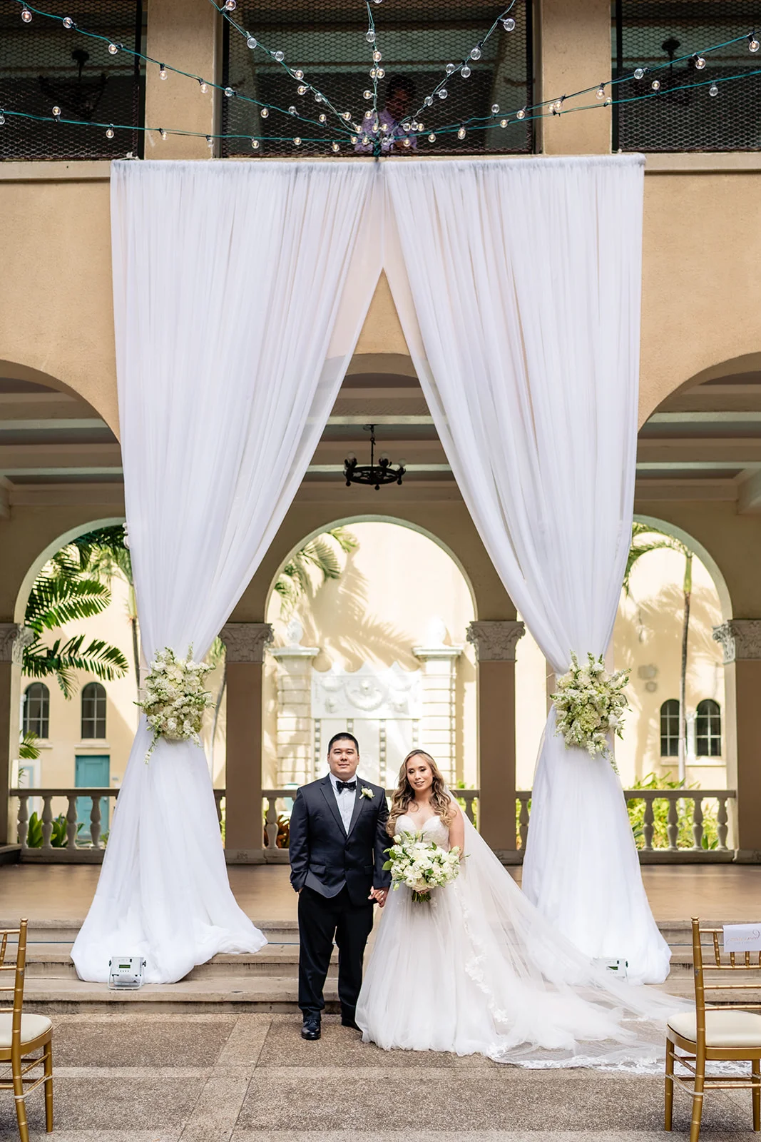 Wedding couple at Cafe Julia on Oʻahu, standing under elegant white drapery with floral arrangements and hanging lights.