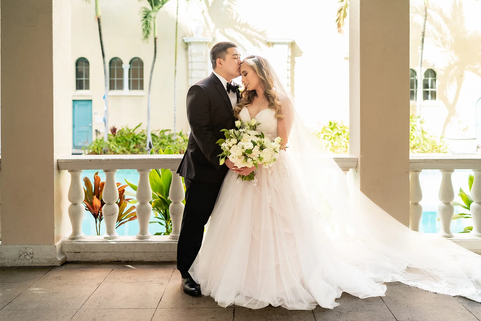 Bride in elegant white gown and groom in black tuxedo with luxury white floral bouquet at Cafe Julia, Oʻahu wedding setting.