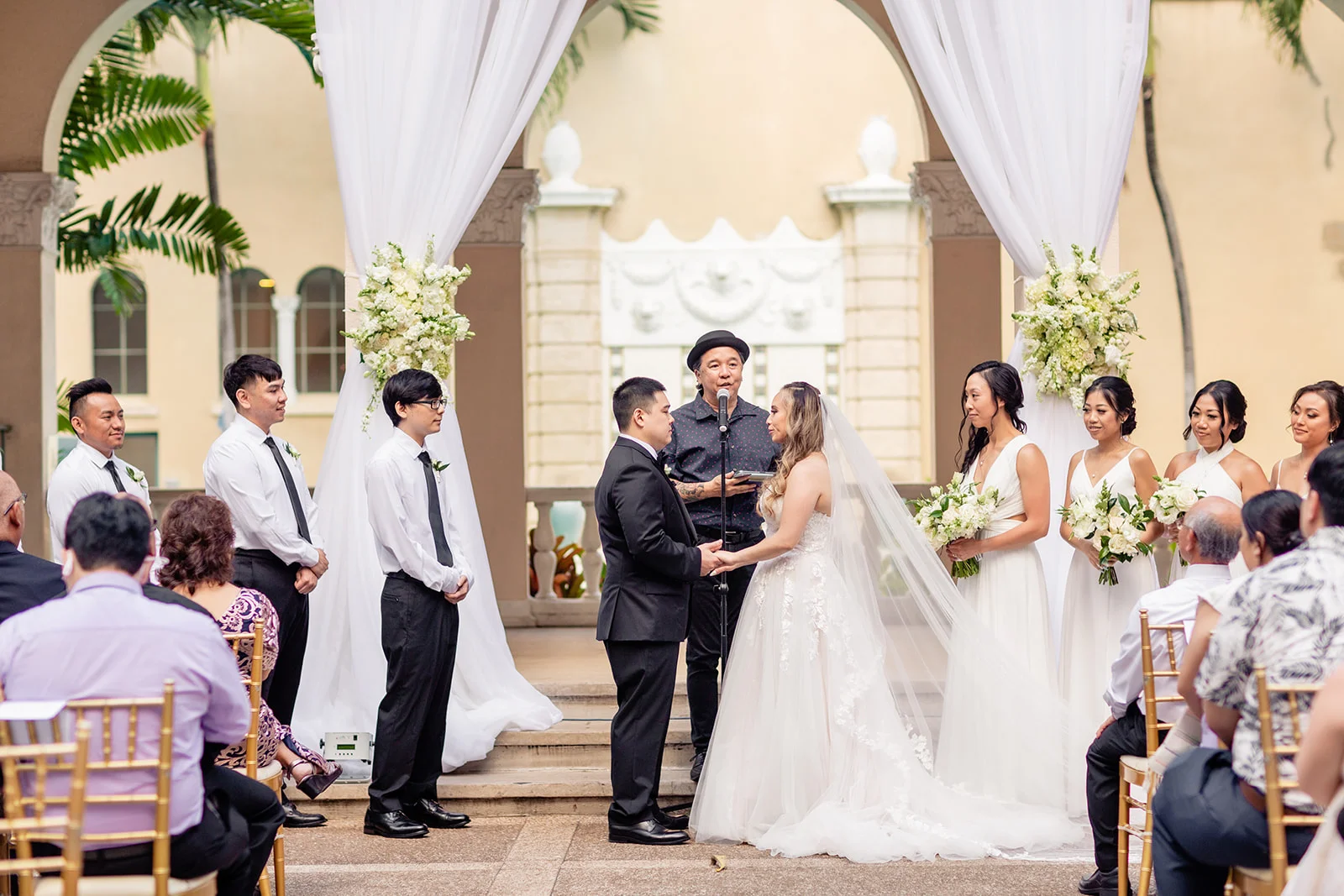 Elegant wedding ceremony at Cafe Julia, Oʻahu, with couple under white floral archway, bridesmaids in white dresses holding bouquets.
