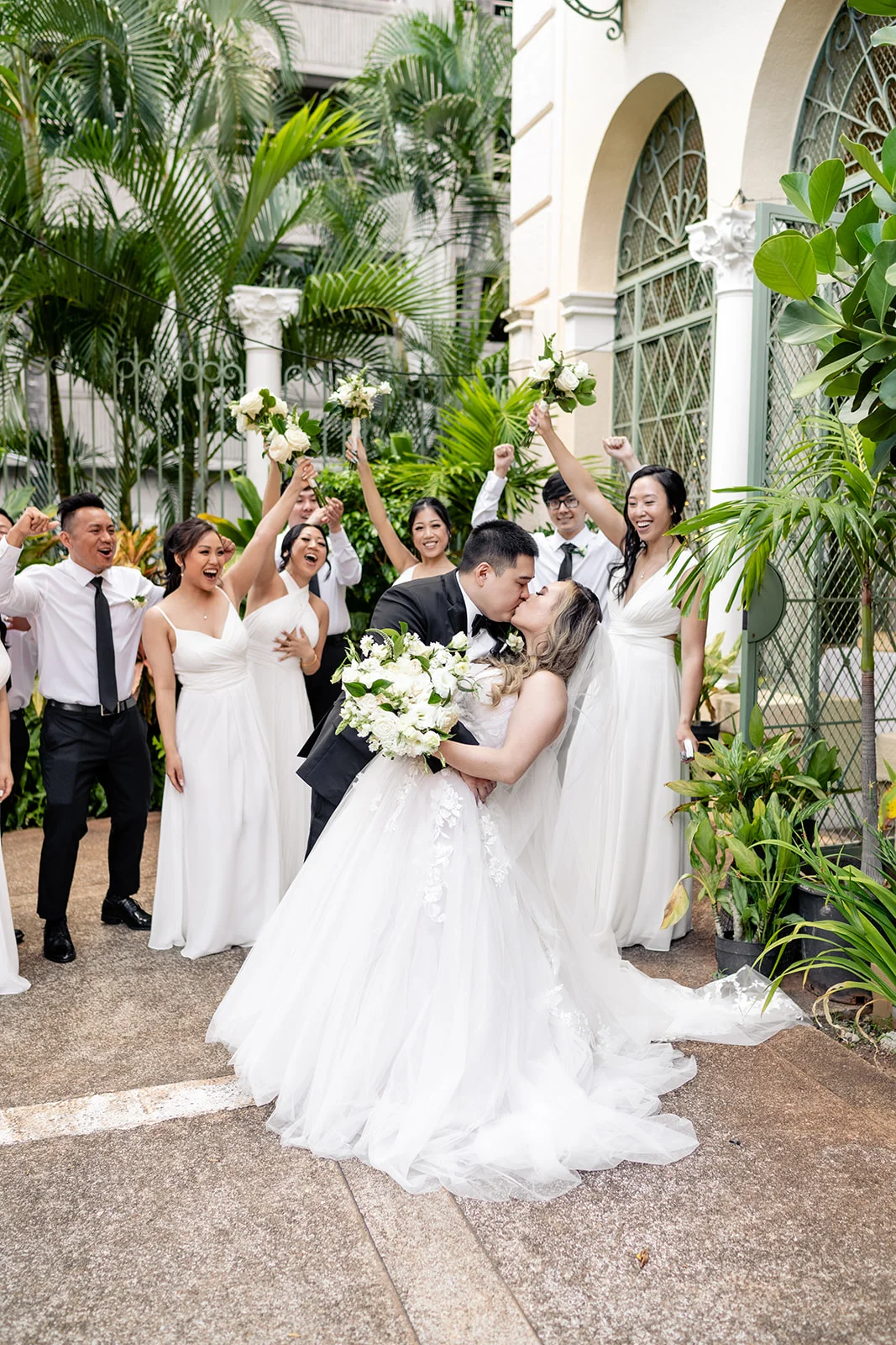 Bride and groom kissing at Cafe Julia, surrounded by cheering bridal party with lush greenery and elegant white bouquets, Oʻahu.