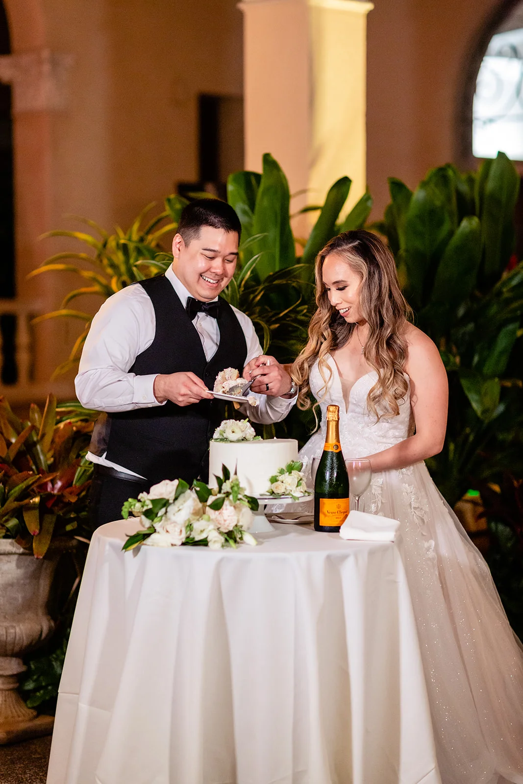 Couple cutting a white wedding cake with floral decorations and champagne at Cafe Julia on Oʻahu.