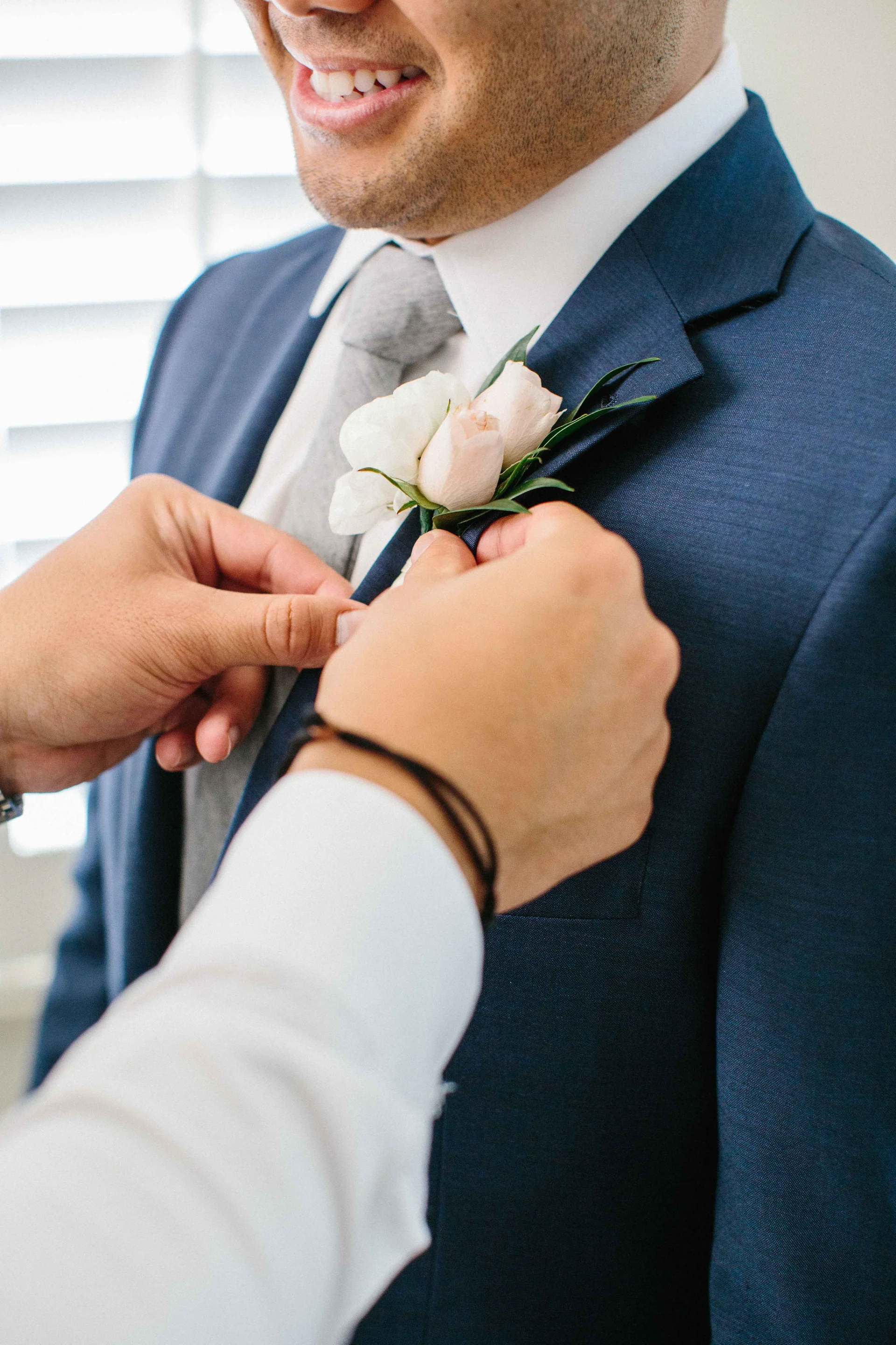 Groom wearing a navy suit with a pale pink rose boutonniere, being adjusted by another person at a wedding in Oʻahu.