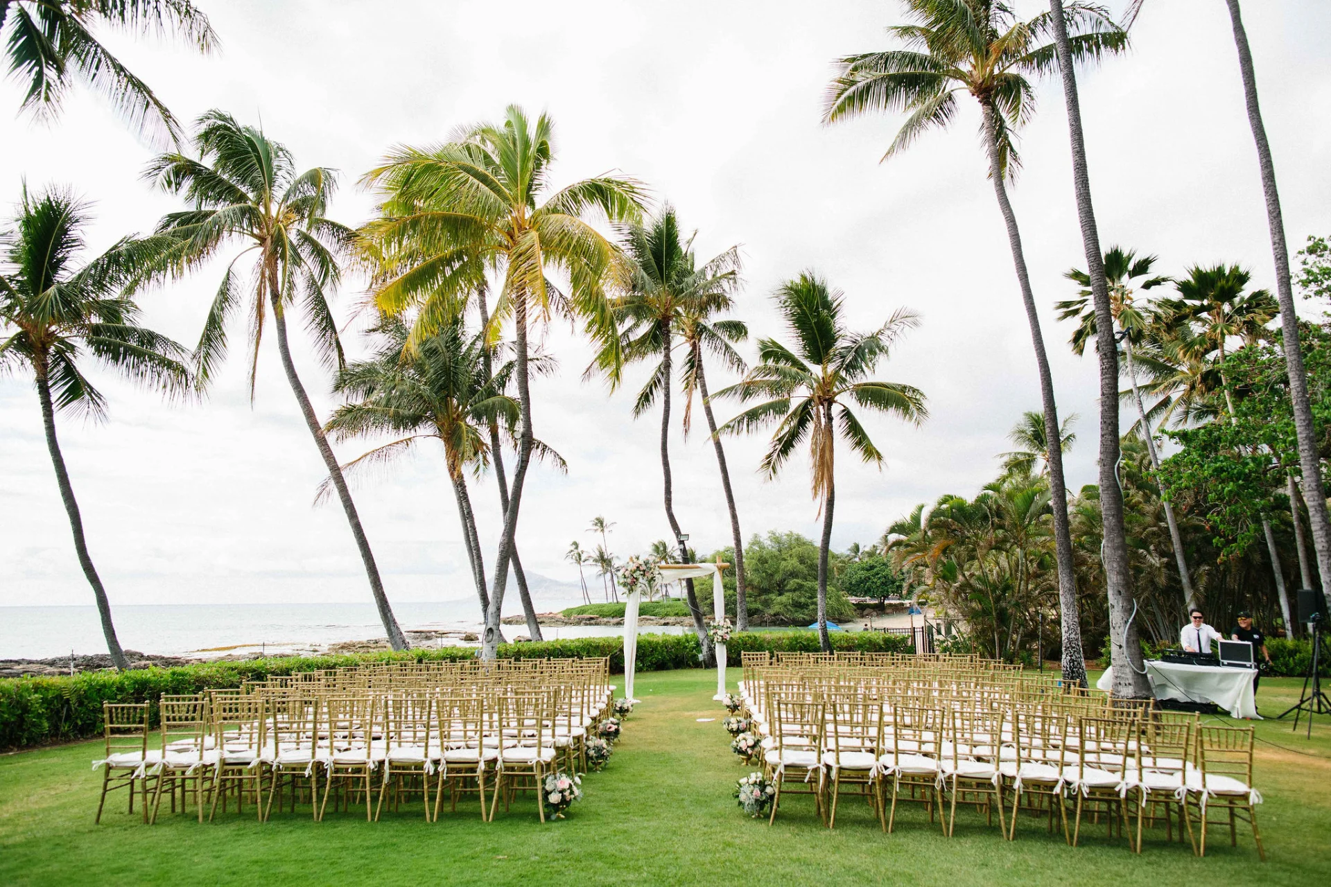 Beachfront wedding setup at Lanikūhonua Cultural Institute with rows of gold chiavari chairs and floral decorations under palm trees.