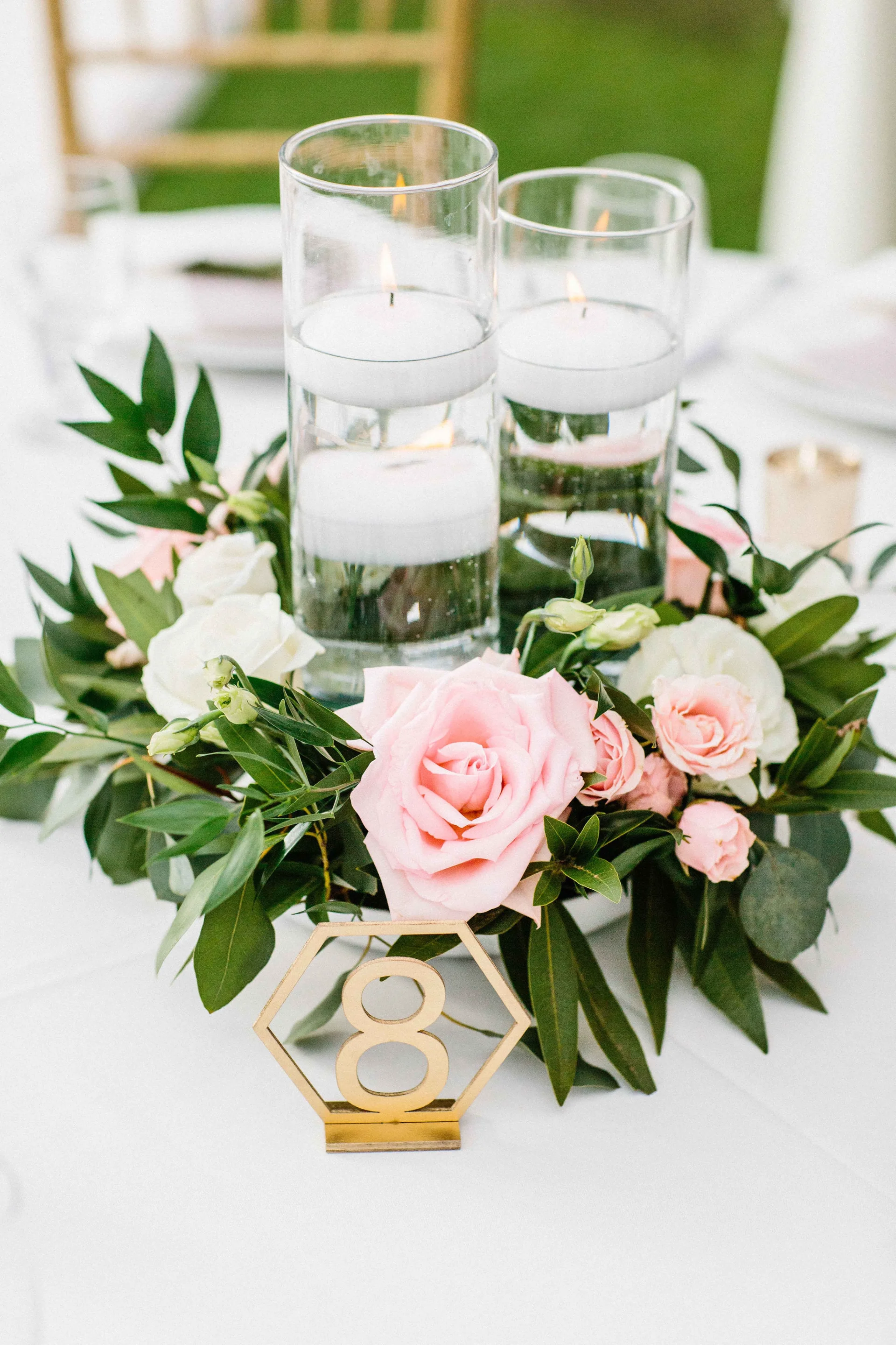 Elegant wedding centerpiece with pink and white roses, greenery, floating candles, and gold table number at Lanikūhonua, Oʻahu.